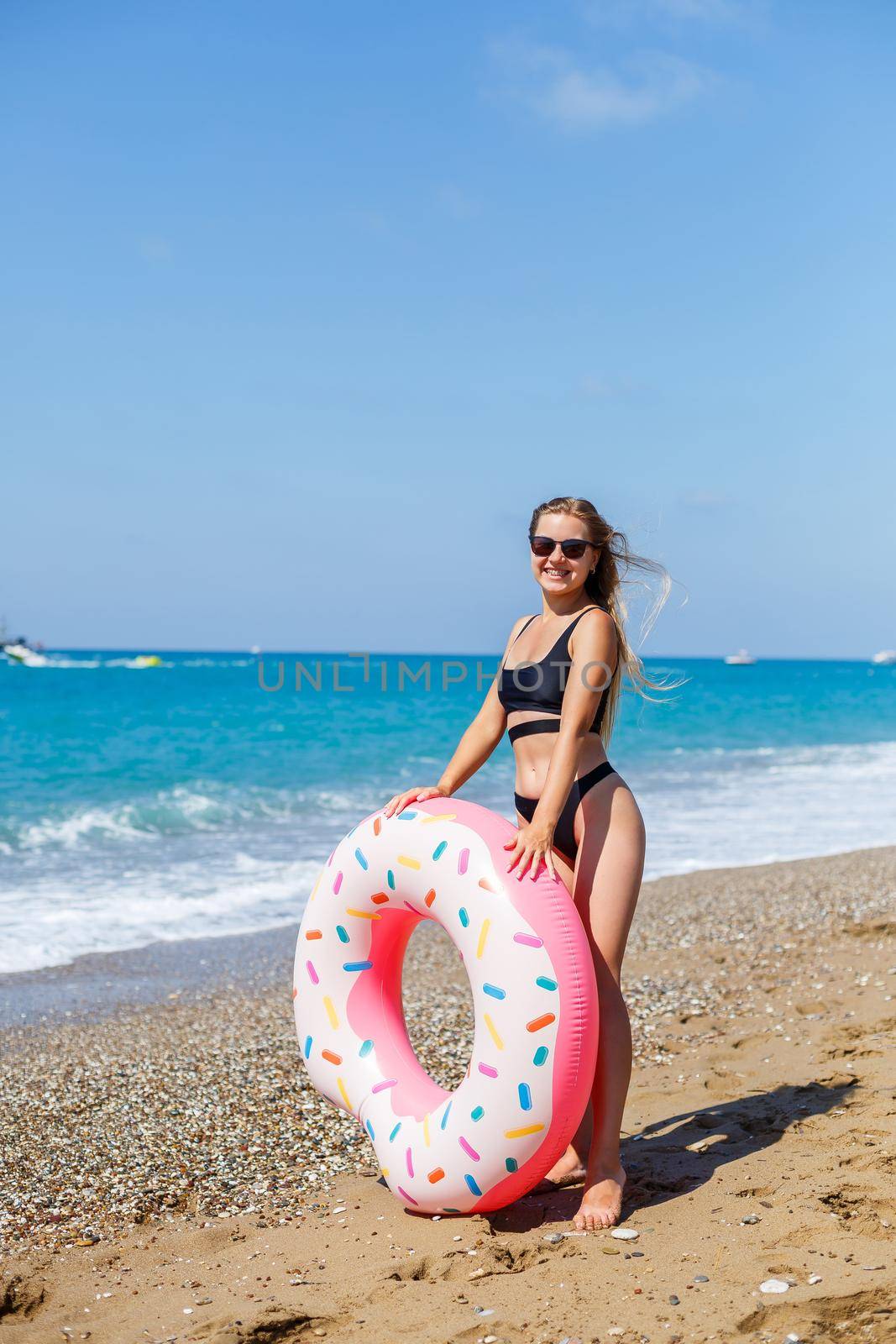 A slender woman in a stylish black swimsuit and sunglasses holds an inflatable circle donut in her hand. Beautiful happy girl posing and having fun in the sun. Vacation. travel by sea