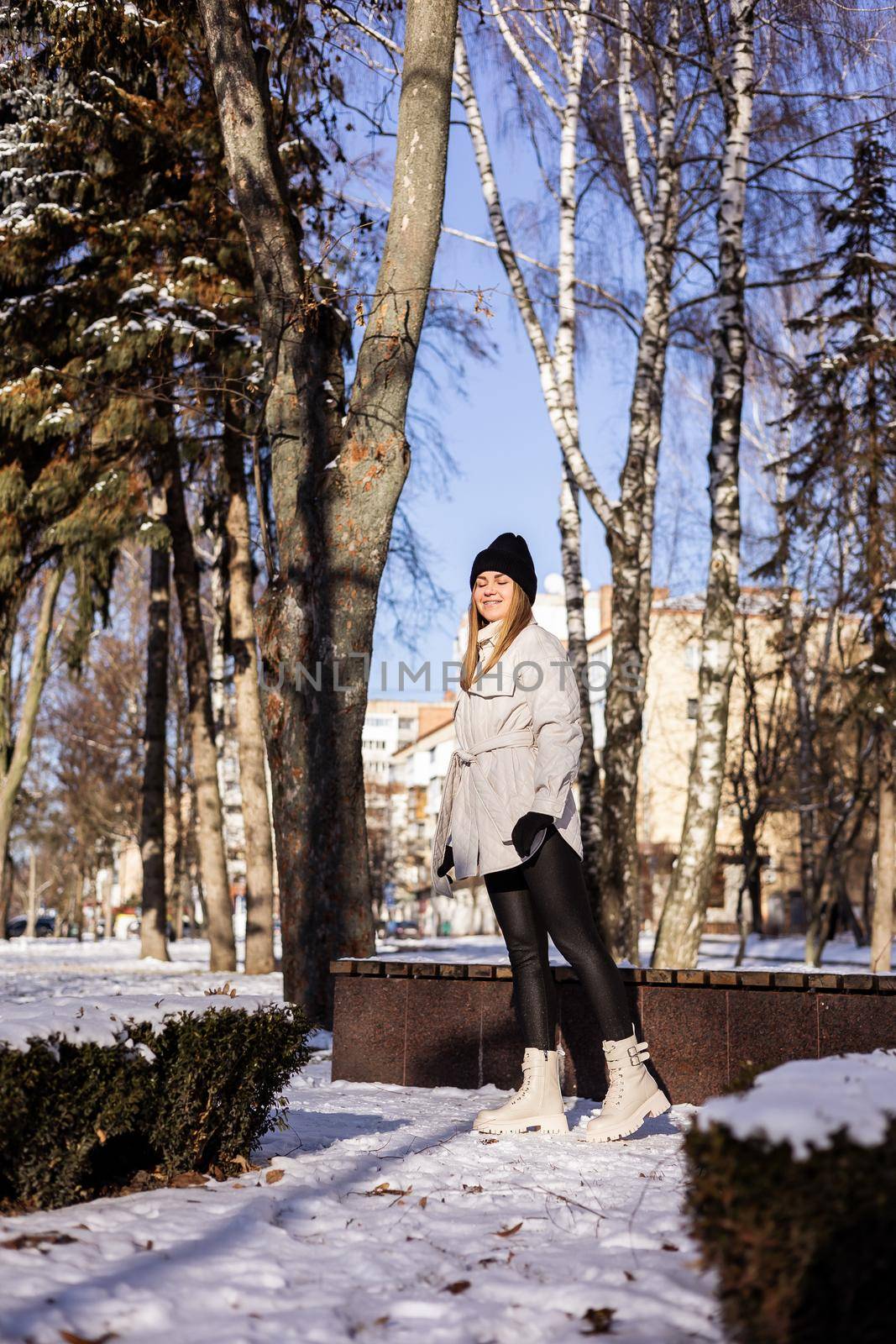 A beautiful young woman in a white jacket and white leather boots walks in a snowy park