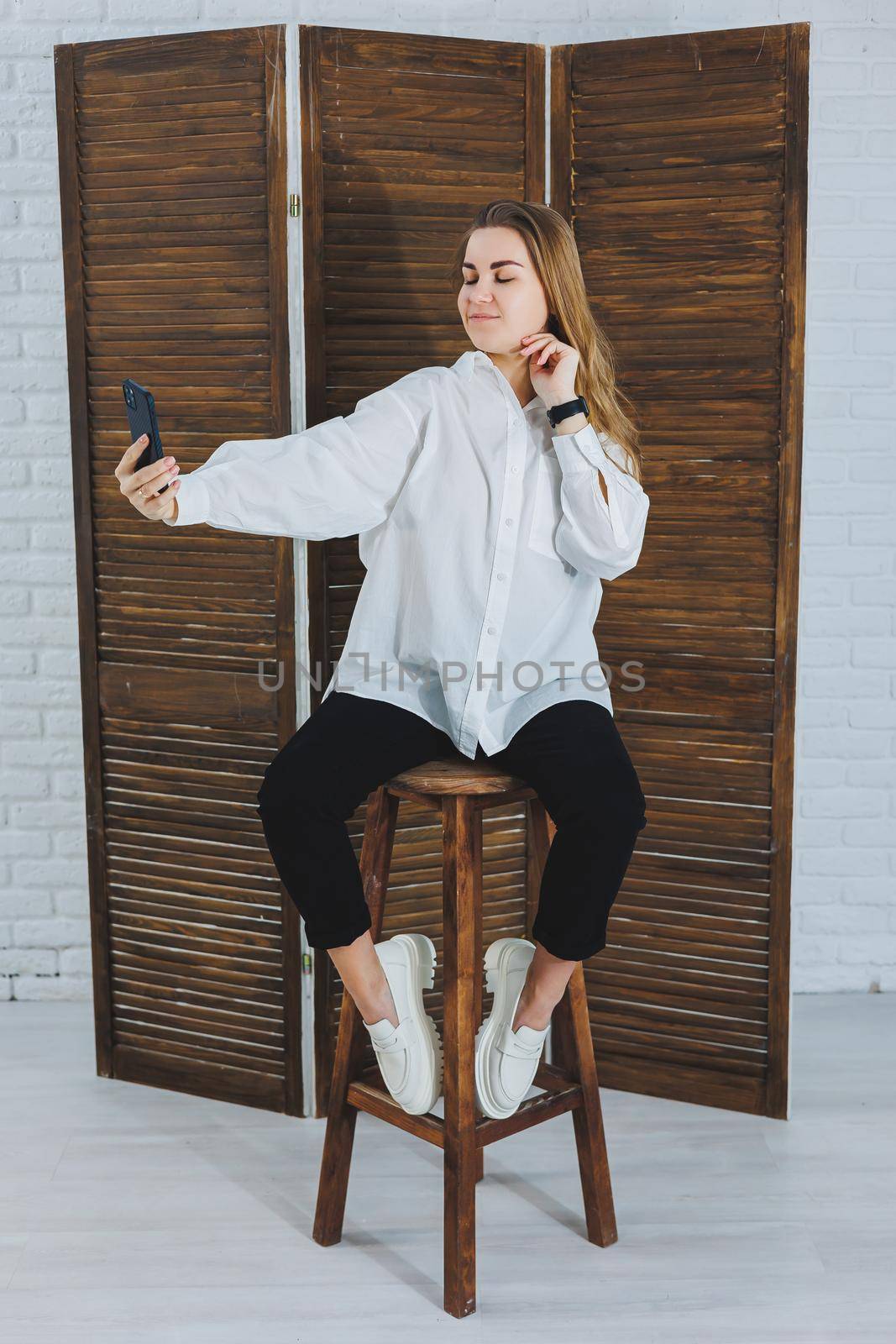 Blonde woman with long hair in a white shirt and black pants posing at the phone camera, holding the phone and taking a selfie on an isolated background.