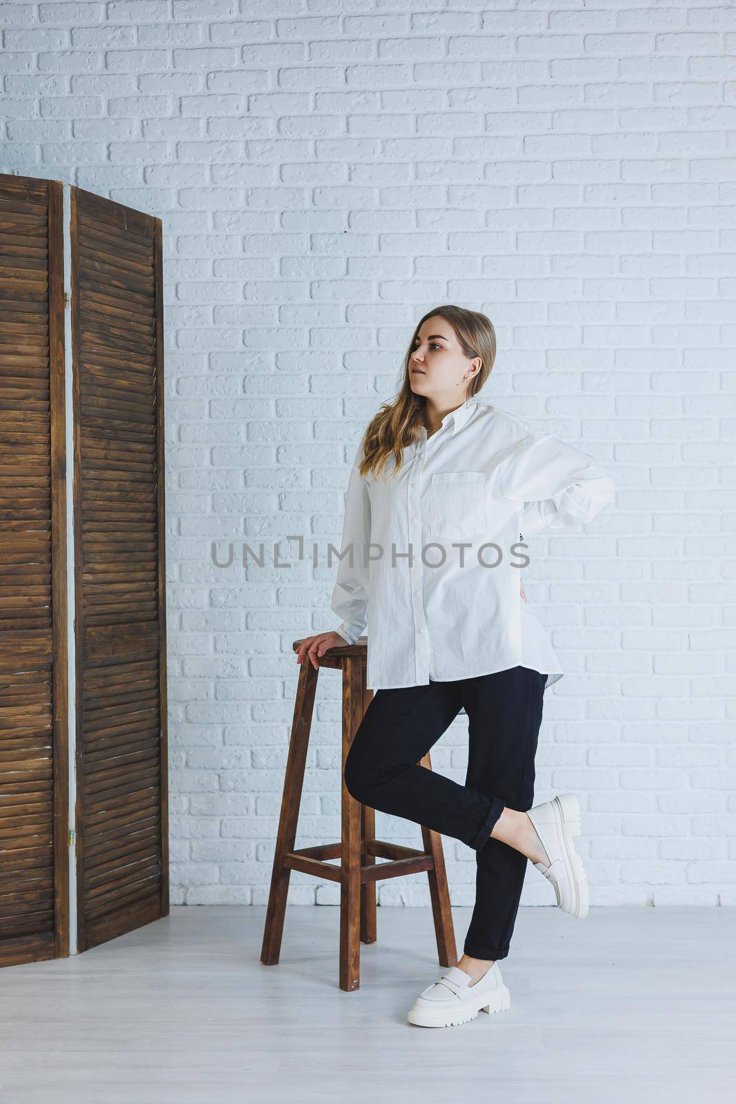 A young woman in black pants and a white shirt against a white brick wall, a stylish modern girl in white leather loafers