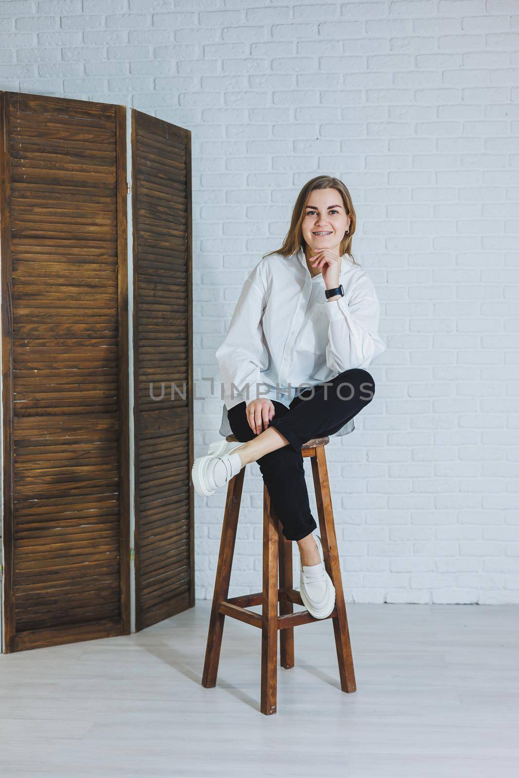 A beautiful woman is wearing a white cotton shirt and black pants. Cheerful emotional girl sits on a wooden chair against a white brick wall.