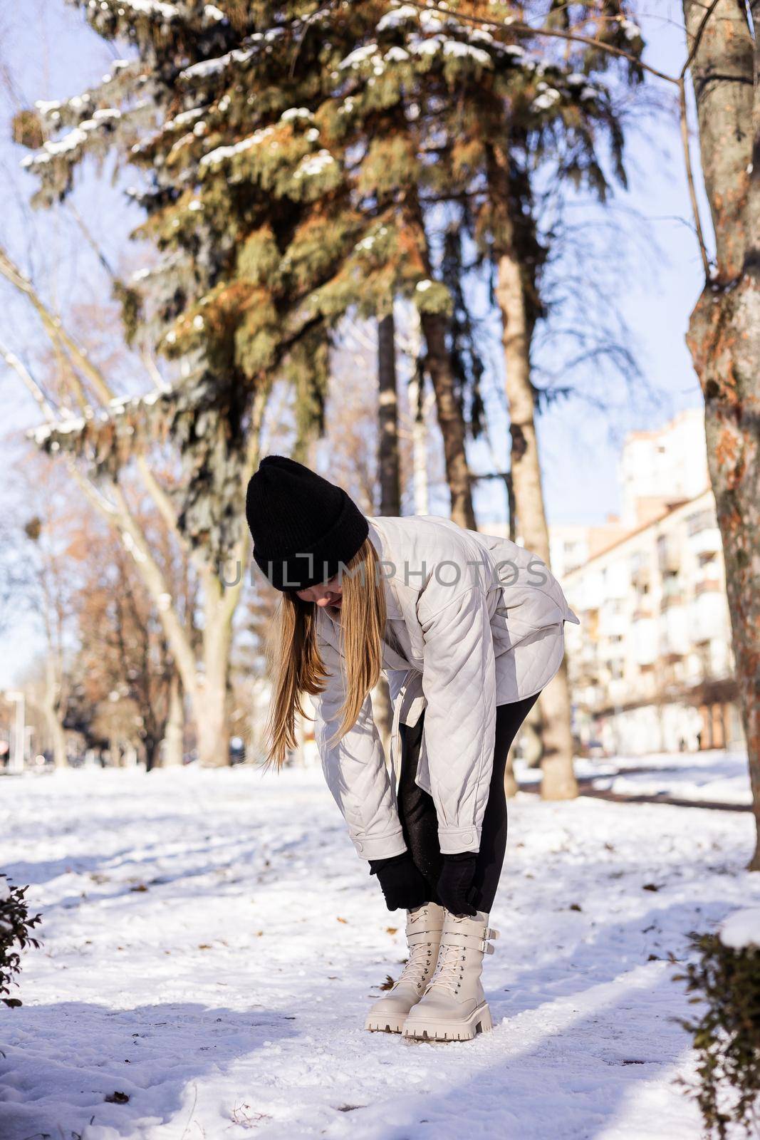 A beautiful young woman in a white jacket and white leather boots walks in a snowy park