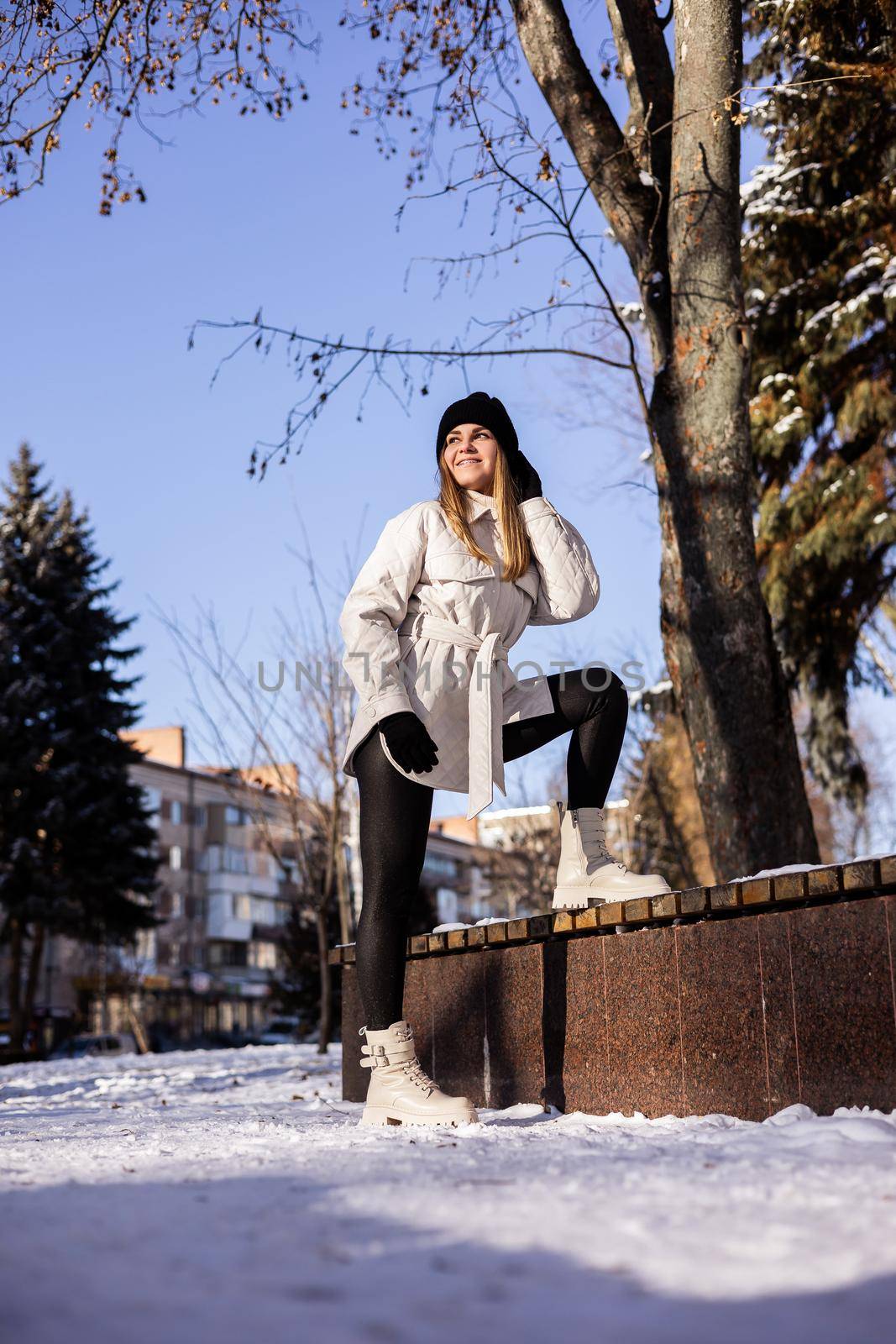 A beautiful young woman in a white jacket and white leather boots walks in a snowy park