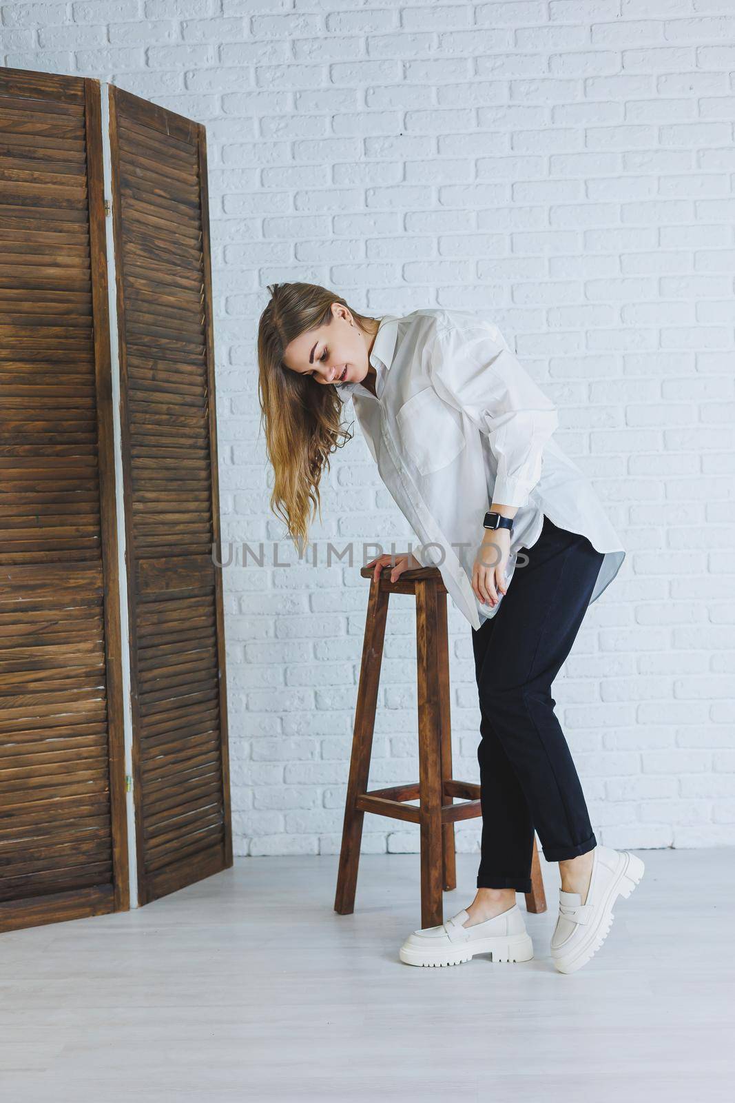 A young woman in black pants and a white shirt against a white brick wall, a stylish modern girl in white leather loafers