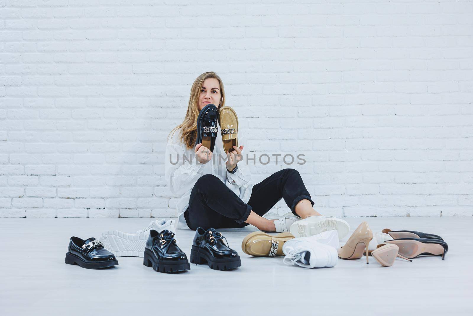 Happy beautiful young woman with a smile in a fashionable outfit with a white shirt and black trousers sits on the floor among the shoes and chooses a new pair of shoes for herself