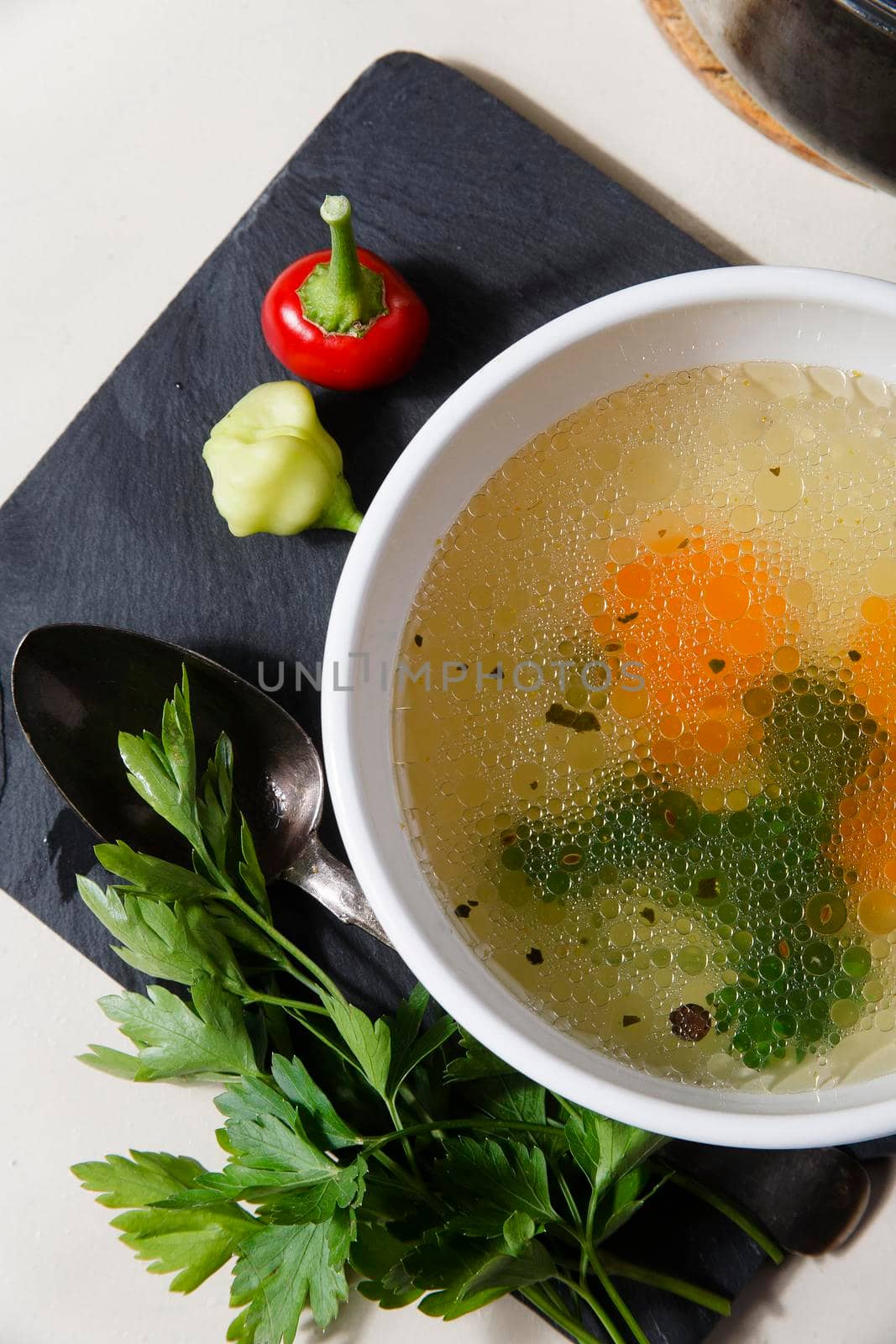 A bowl with fresh home made chiken broth with carrot, parsley and red chili pepper on black cutting board on white table, top view, close up.