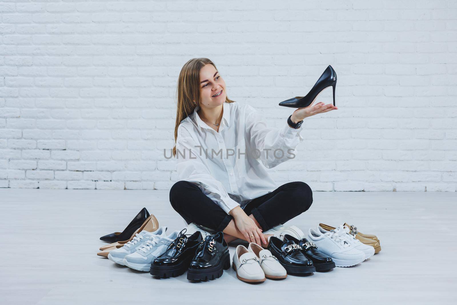 Stylish blonde choosing shoes while sitting on the floor. Side view of a lot of women's shoes on the floor, a choice of stylish shoes.