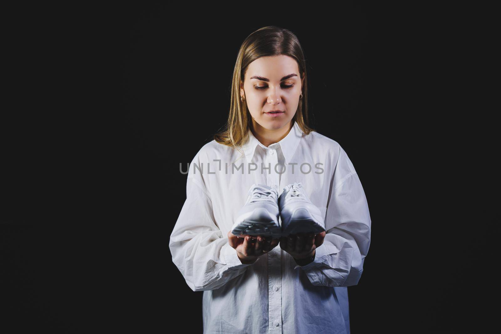 A fair-haired woman in a white shirt on a black background holds white sneakers made of genuine leather in her hands