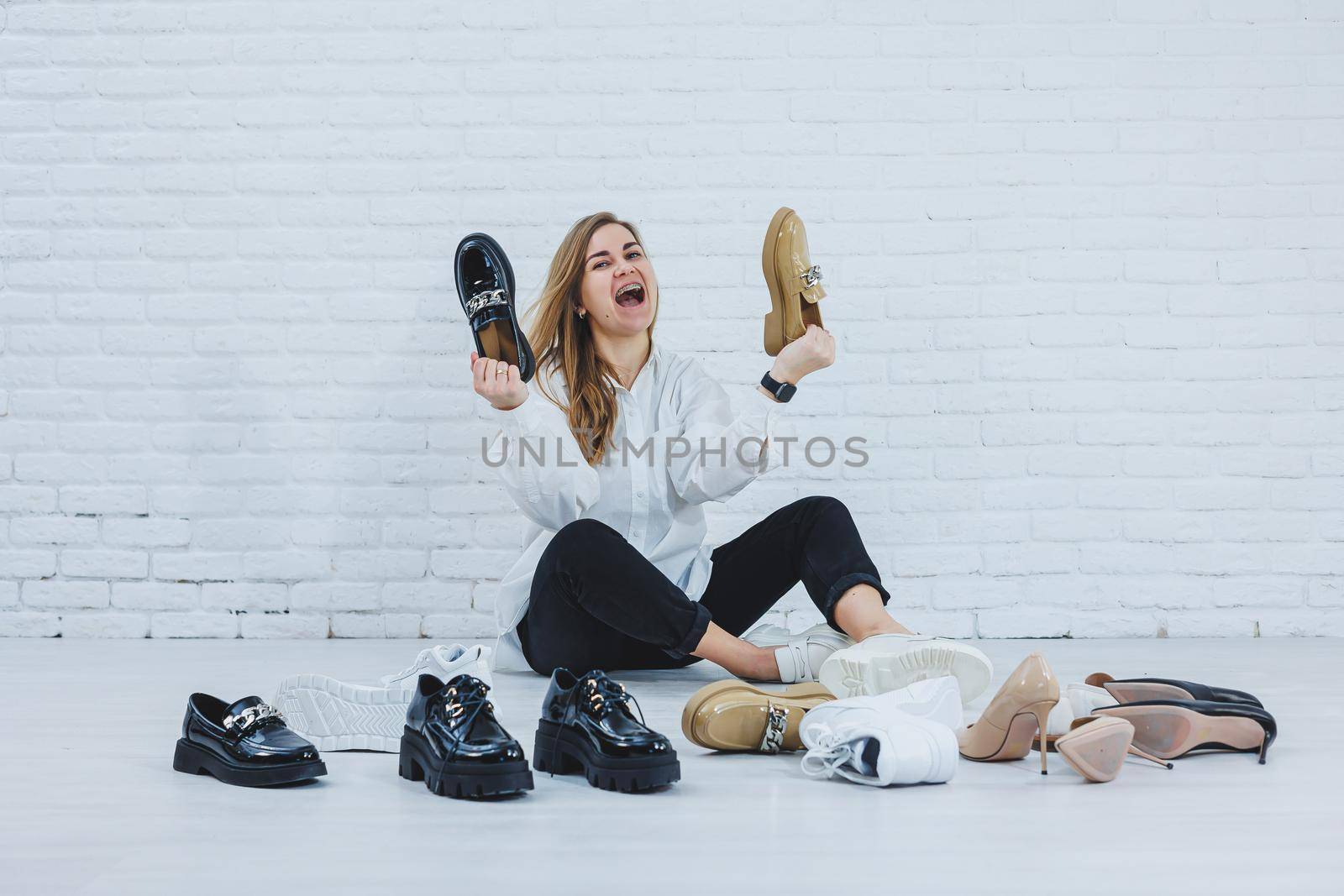 Happy beautiful young woman with a smile in a fashionable outfit with a white shirt and black trousers sits on the floor among the shoes and chooses a new pair of shoes for herself