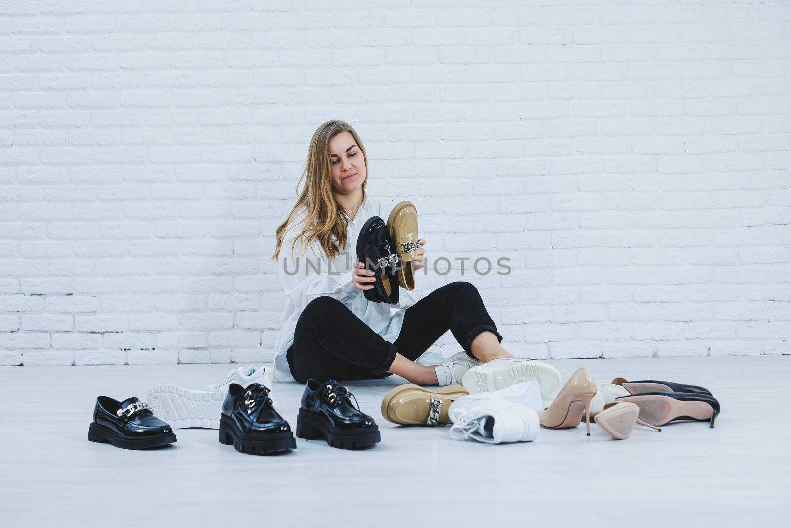 Happy beautiful young woman with a smile in a fashionable outfit with a white shirt and black trousers sits on the floor among the shoes and chooses a new pair of shoes for herself