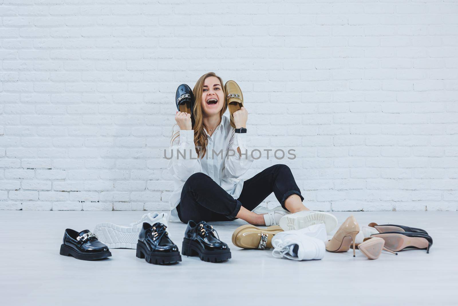 Happy beautiful young woman with a smile in a fashionable outfit with a white shirt and black trousers sits on the floor among the shoes and chooses a new pair of shoes for herself