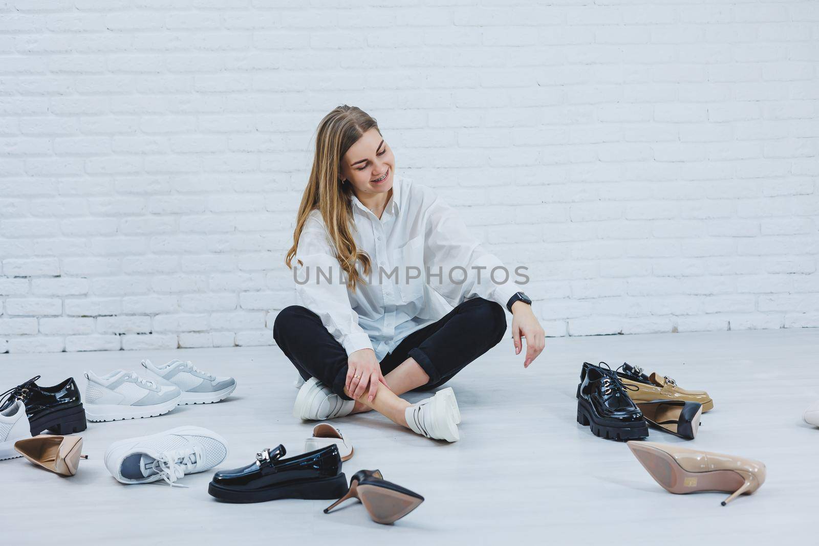 A young woman sits on the floor among shoes and chooses new shoes for herself. Lady in a white shirt and black trousers. Woman in white shoes.