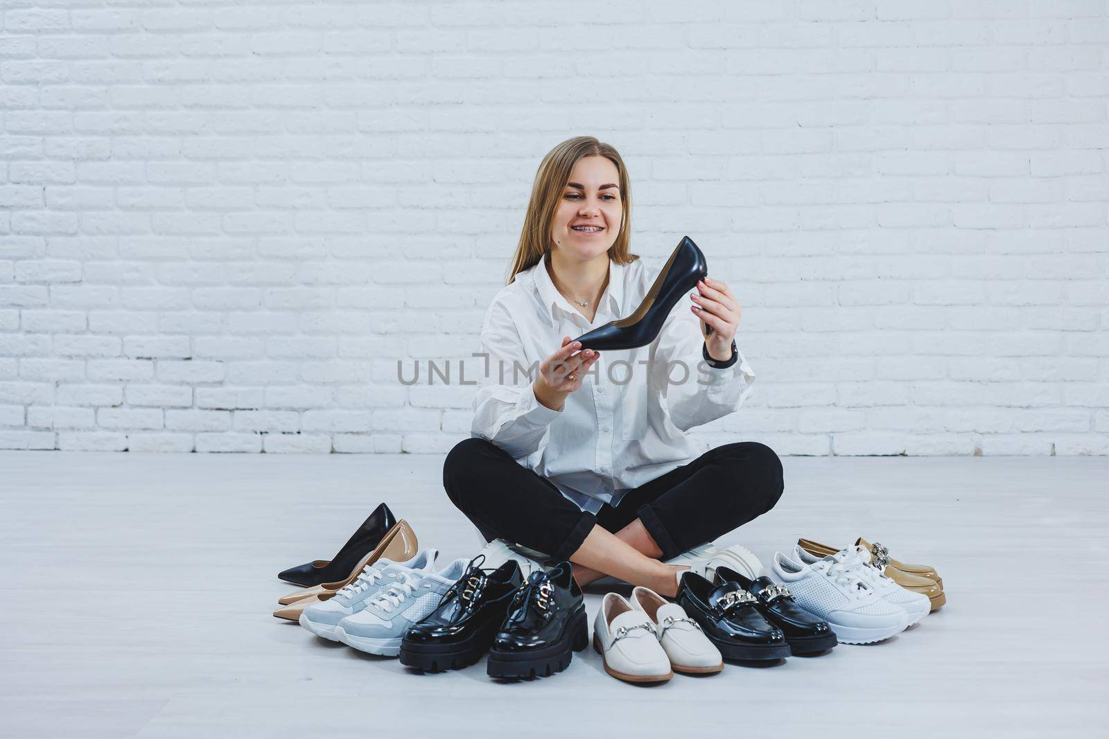 Stylish blonde choosing shoes while sitting on the floor. Side view of a lot of women's shoes on the floor, a choice of stylish shoes.