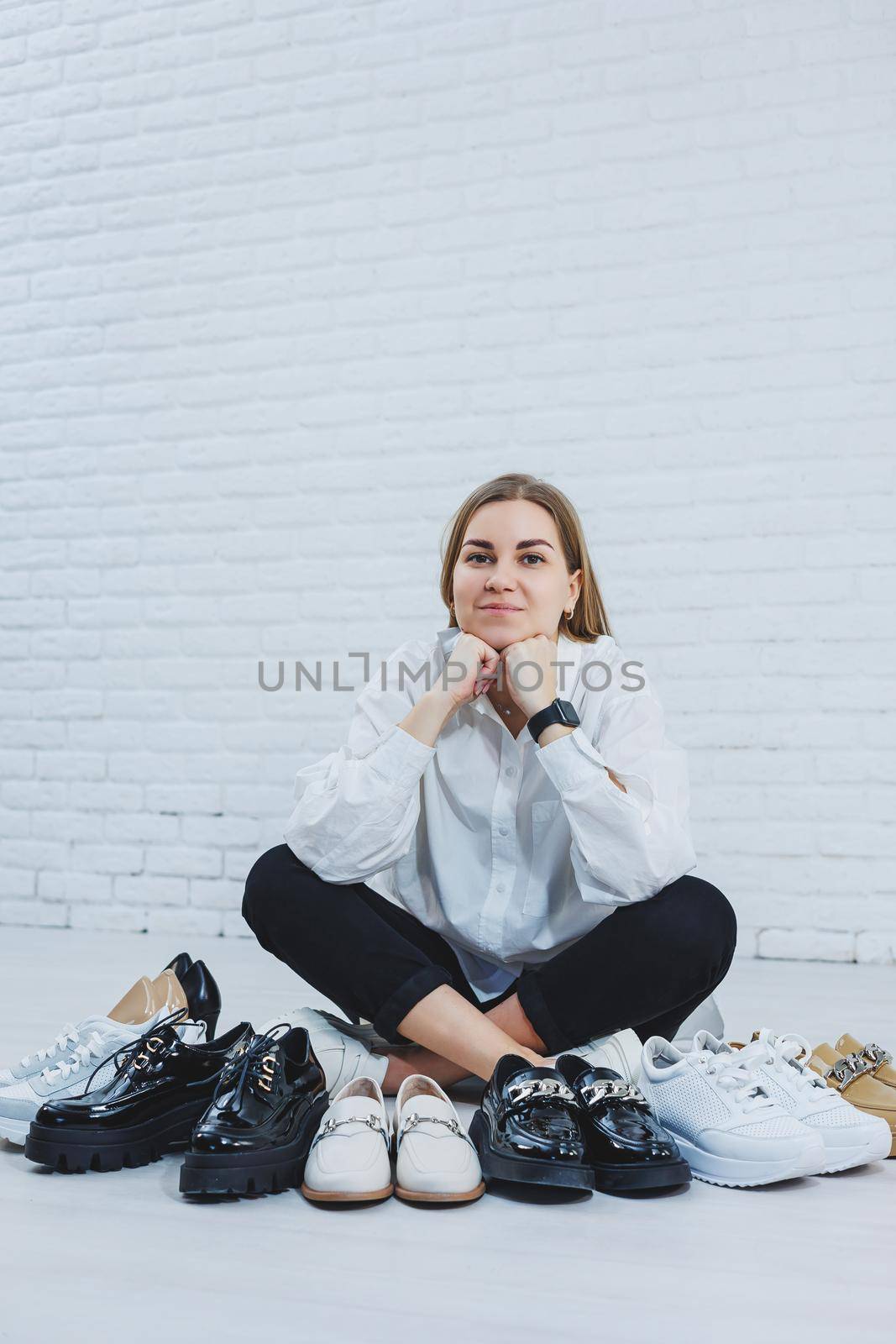 Happy beautiful young woman with a smile in a fashionable outfit with a white shirt and black trousers sits on the floor among the shoes and chooses a new pair of shoes for herself