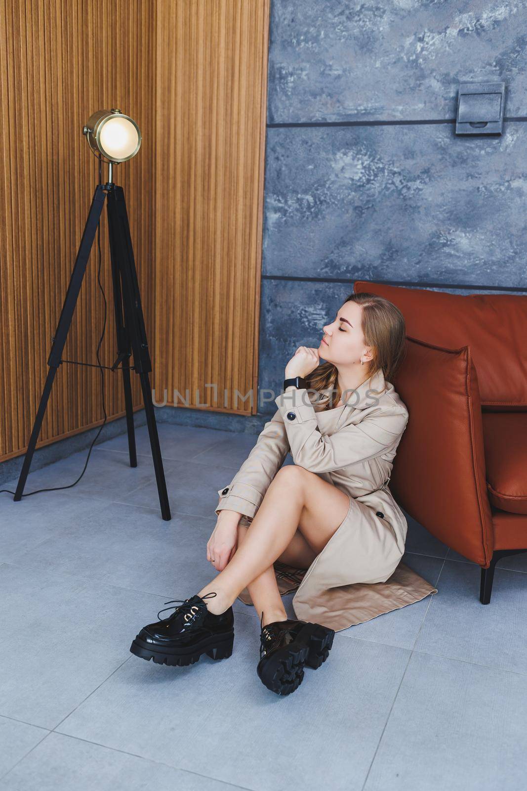 Young beautiful woman sits on the floor in a beige spring raincoat and modern stylish black shoes. Stylish woman of European appearance with long blond hair