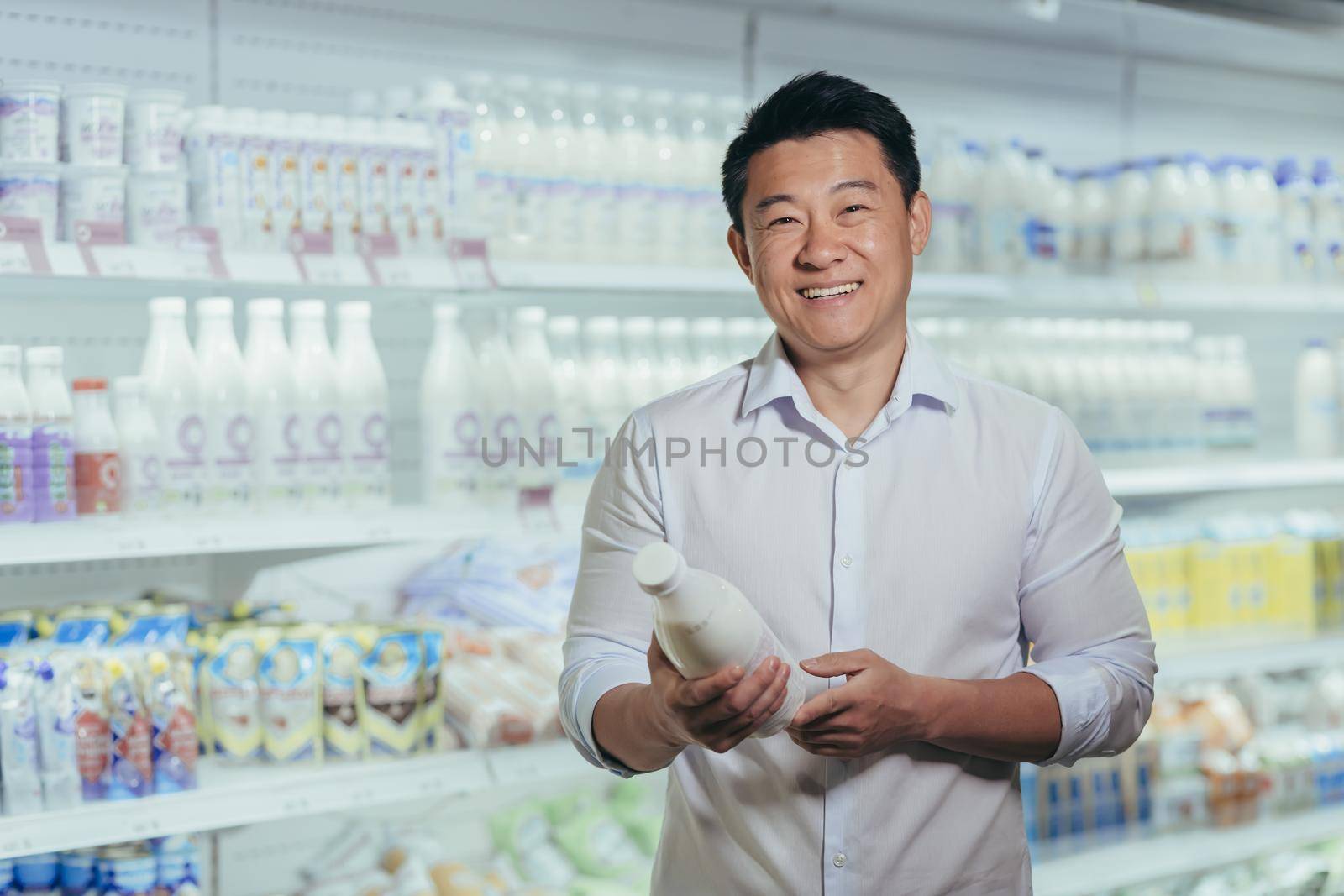 Portrait of happy and satisfied asian shopper in supermarket in dairy department, man holding bottle of milk smiling and looking at camera near refrigerators