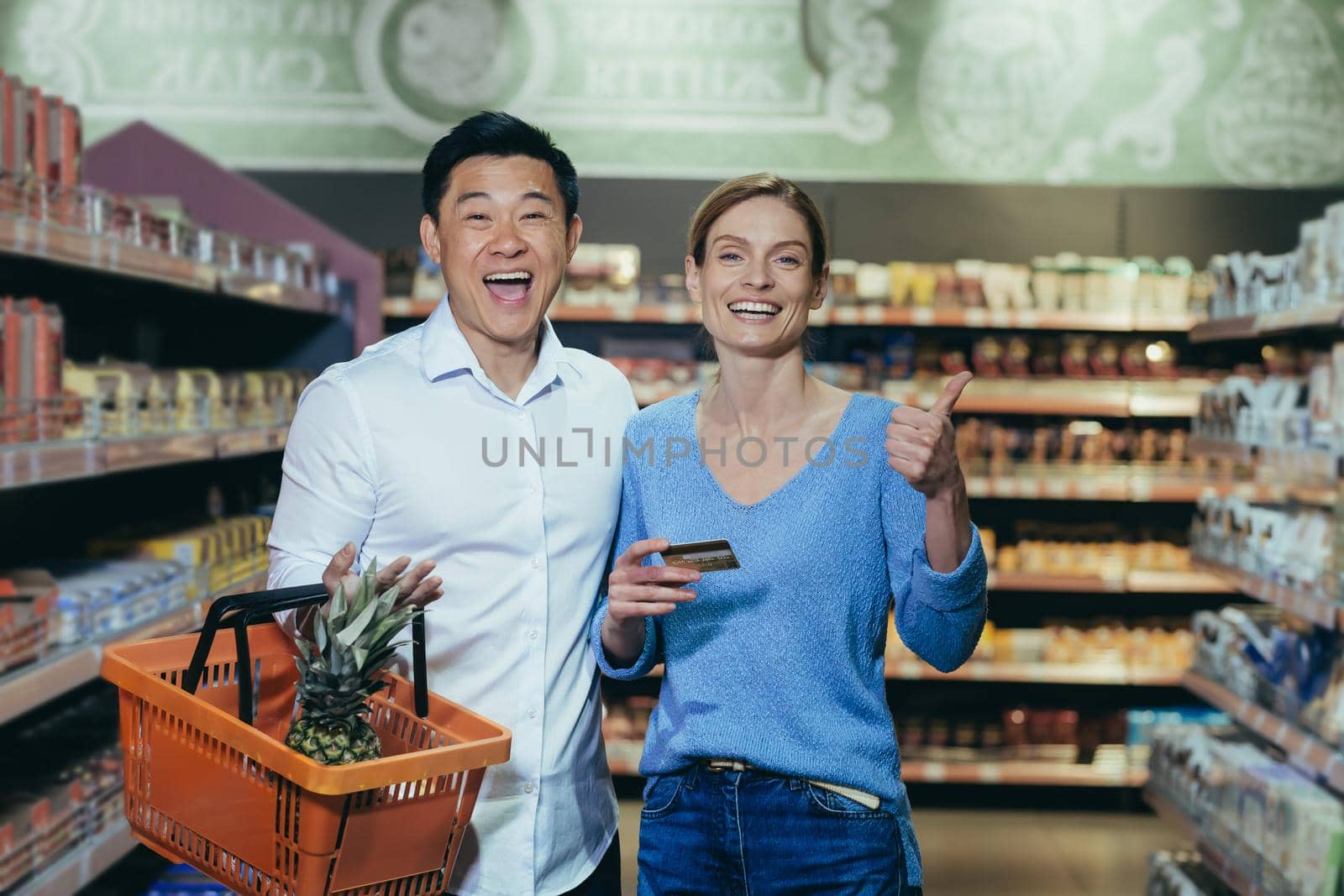 Happy diverse family couple man and woman shoppers in supermarket, looking at camera and smiling happy shopping, holding phone and bank credit card in hand, with shopping basket.