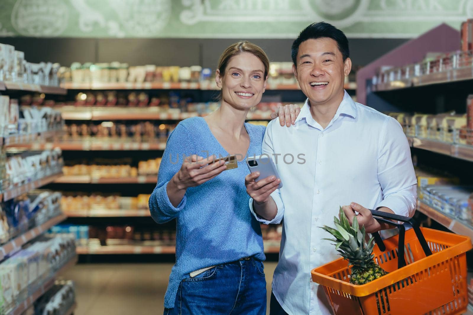 Happy diverse family couple man and woman shoppers in supermarket, looking at camera and smiling happy shopping, holding phone and bank credit card in hand, with shopping basket by voronaman