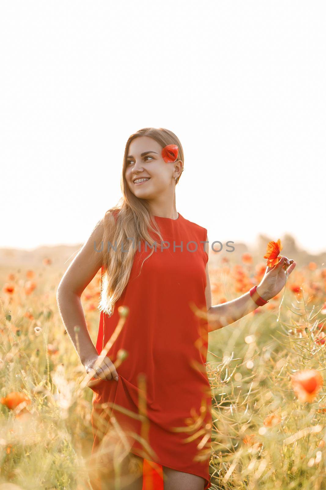 A woman of European appearance with long blonde hair and a red summer dress, she stands in a blooming poppy field