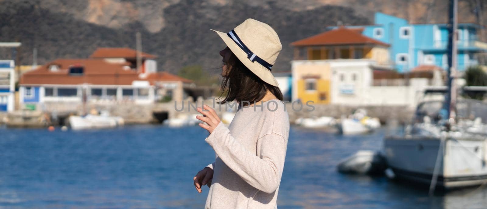 A young girl in a hat looks at the picturesque view with the sea and moored yachts on a sunny day, a backpacker travels and discoveries beautiful places on the coast during her holidays.