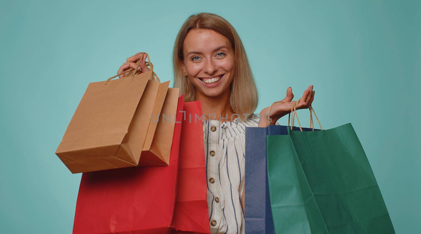 Happy tourist woman showing shopping bags, advertising discounts, smiling looking amazed with low prices, shopping on Black Friday holidays. Young blonde slim thin slender girl on blue background