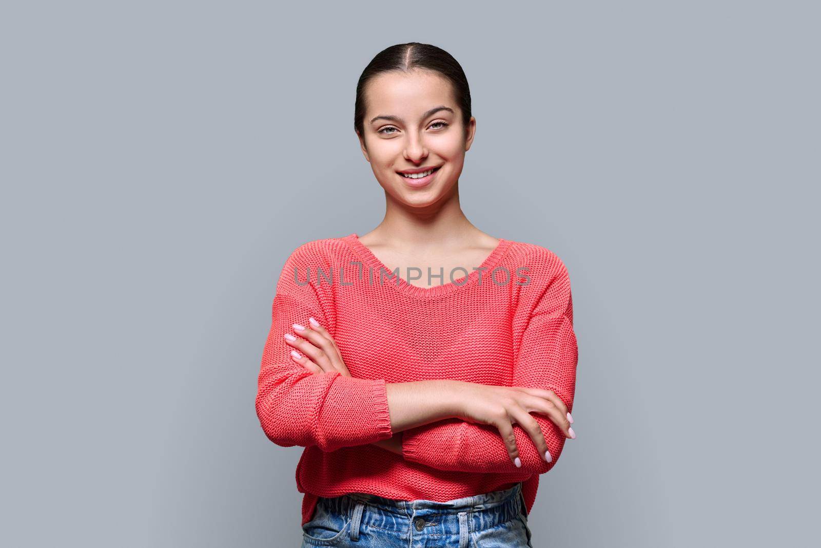 Portrait of teenage smiling female looking at camera on gray color studio background. Confident teen girl with crossed arms in red. Adolescence, high school, youth 15, 16 years old concept