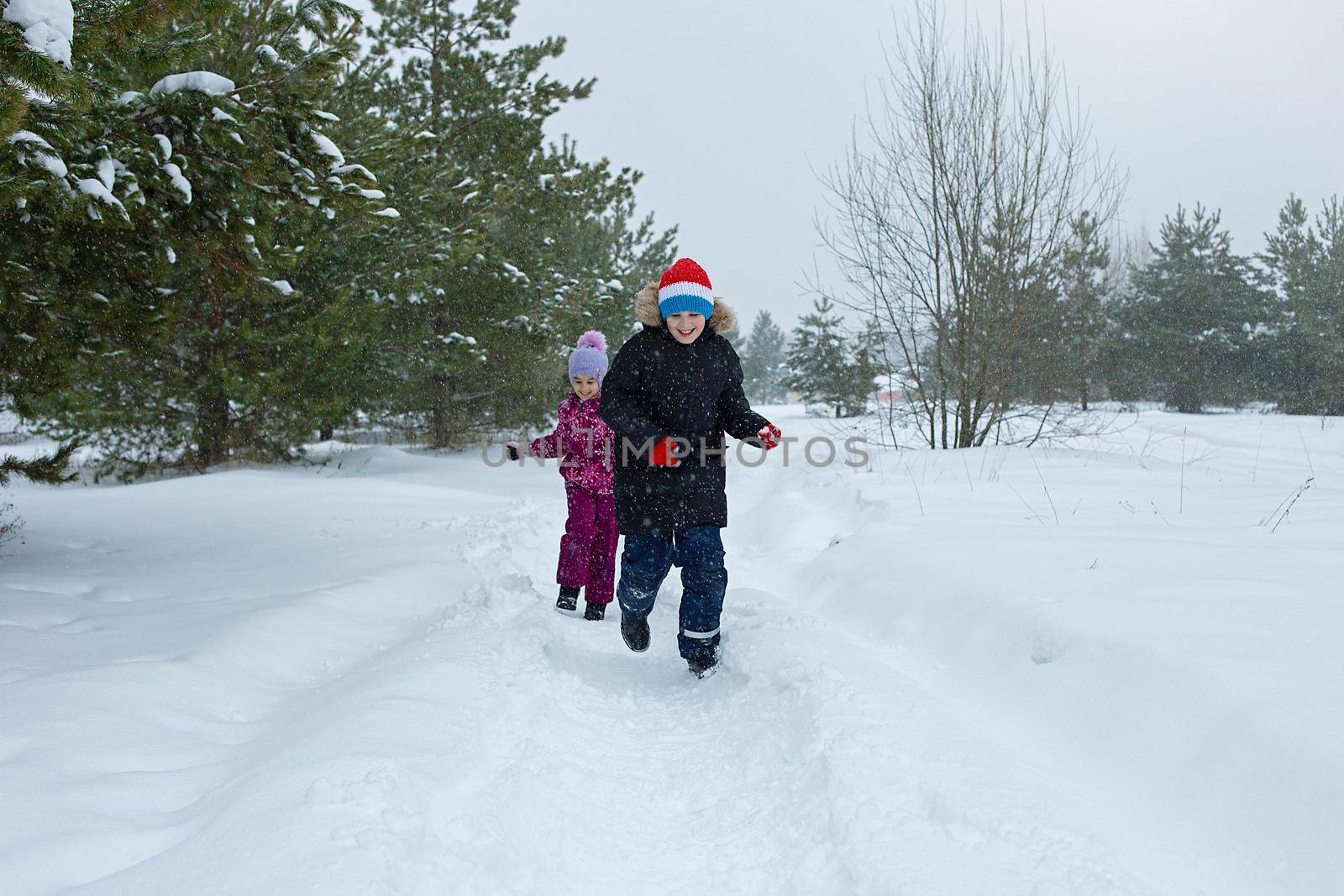 A happy teenager in a winter park runs with a little girl along a path in the snow, next to the pines