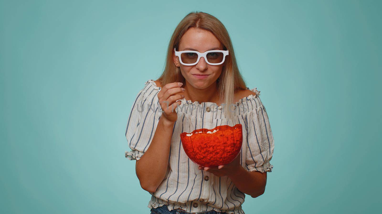 Excited tourist woman in 3D glasses enjoying eating popcorn and watching interesting tv serial sport game, film, online social media movie content. Young girl isolated alone on blue studio background