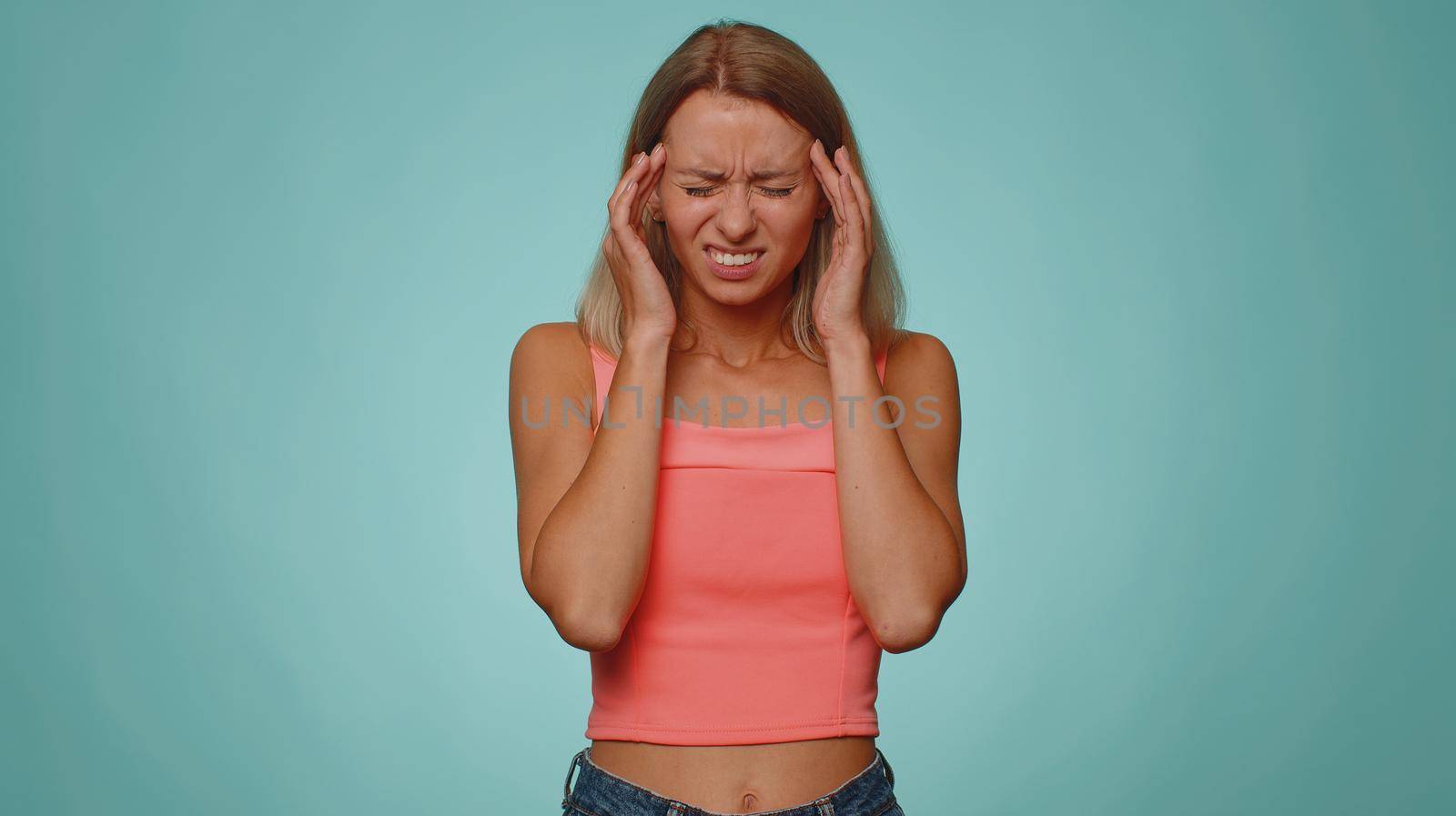 Blonde young adult woman in crop top rubbing temples to cure headache problem, suffering from tension and migaine, stress, grimacing in pain, high blood pressure isolated alone on blue wall background