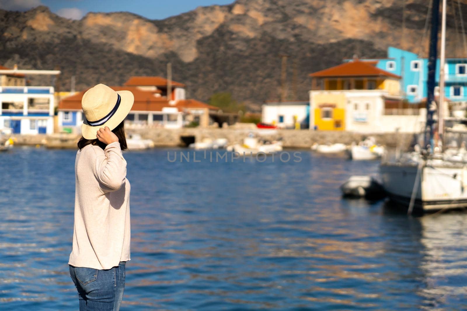 A young girl in a hat looks at the picturesque view with the sea and moored yachts on a sunny day, a backpacker travels and discoveries beautiful places on the coast during her holidays.