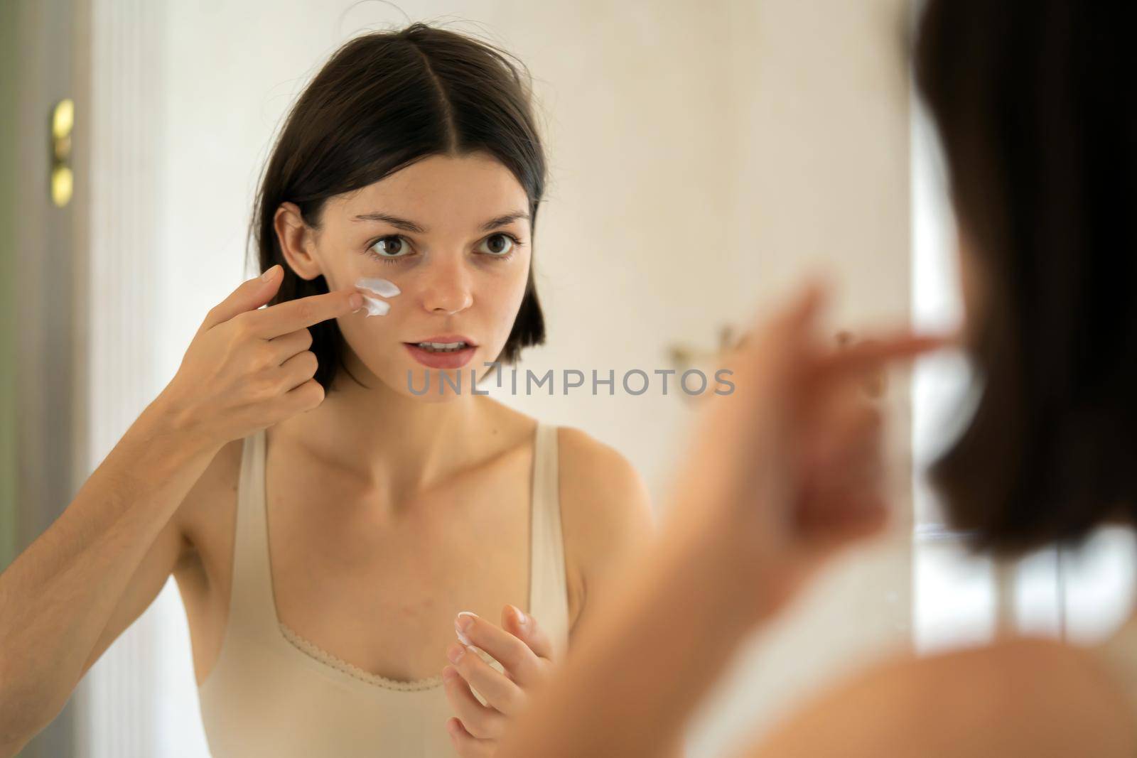 A young pretty girl applies cream stripes on the cheeks, declaring war on dry skin and acne. A woman takes care of the health and hydration of her skin in the bathroom near the mirror.