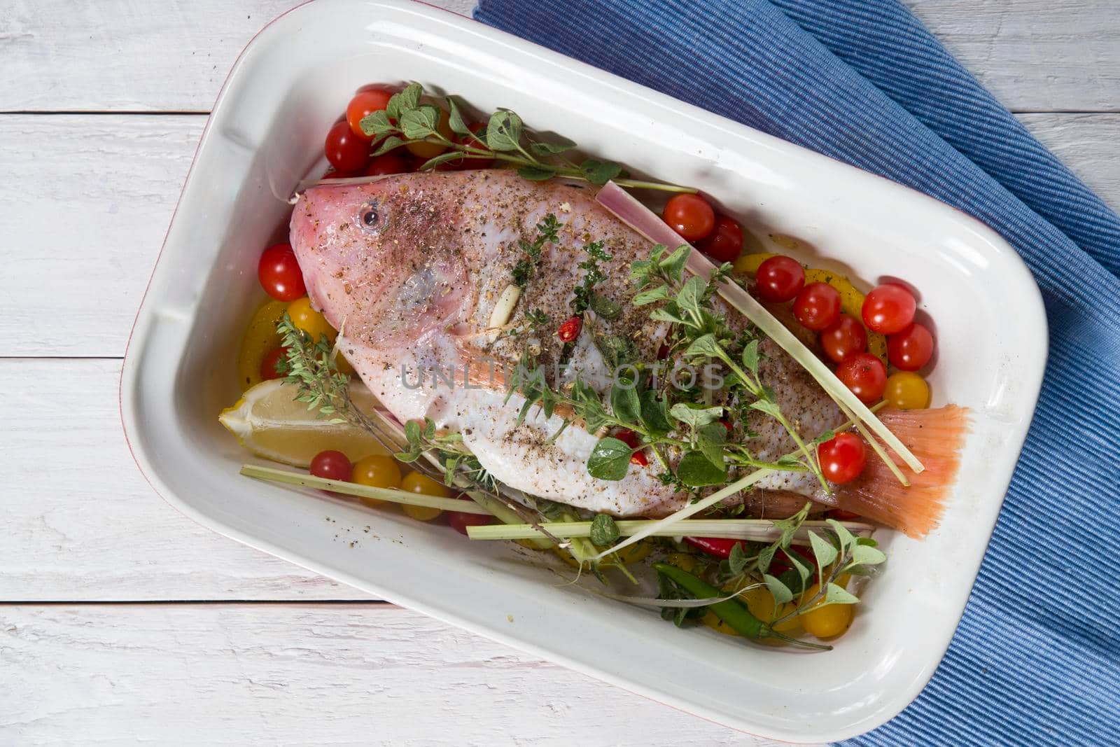 raw pink tilapia fish with vegetables in a baking dish, hands spice on the fish, step by step recipe for white wooden planks,High quality photo