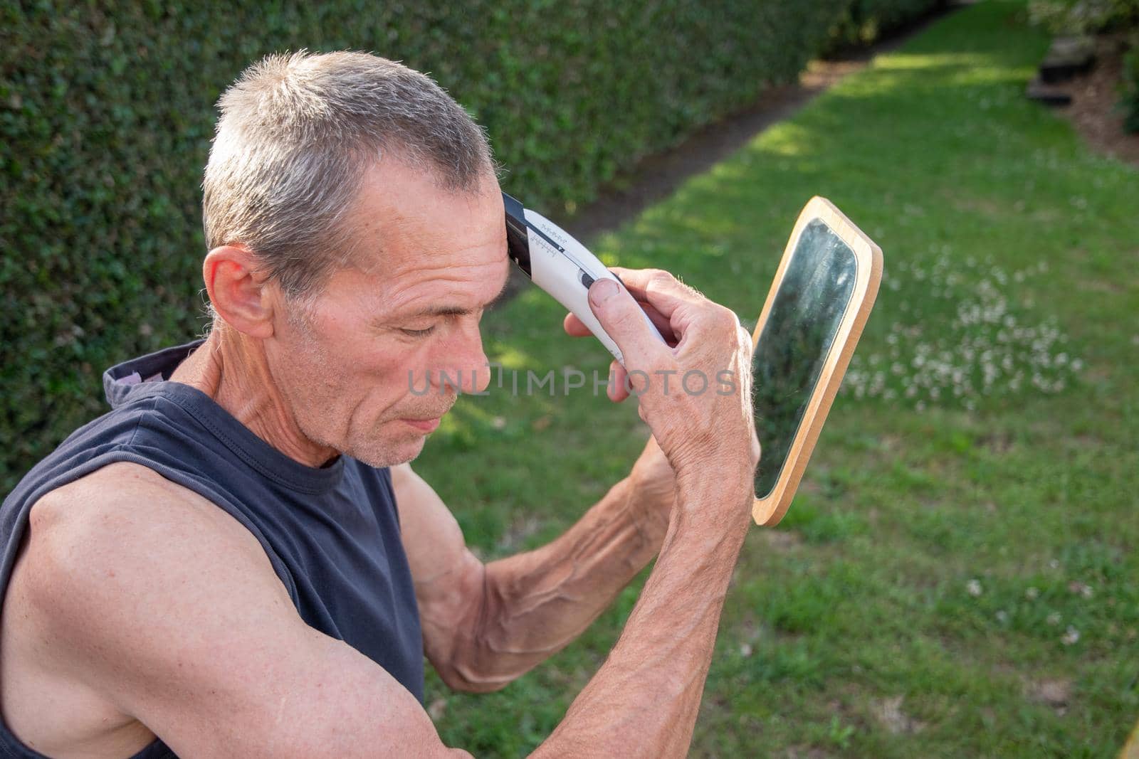 a gray-haired middle-aged man shaves his hair with a clipper in a garden by KaterinaDalemans