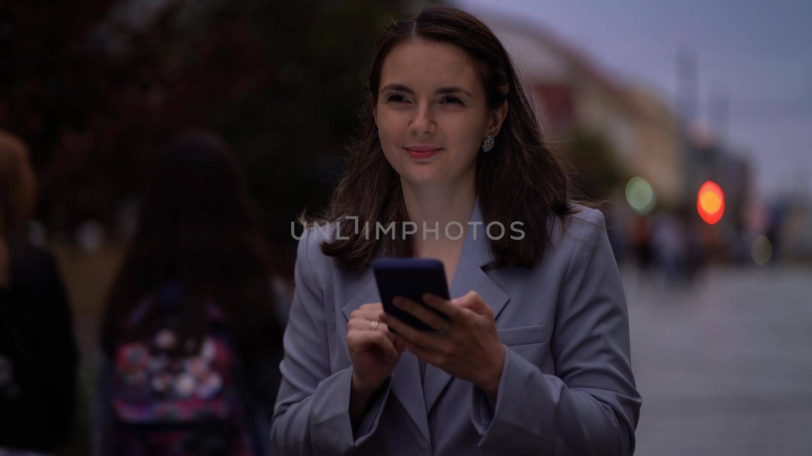 Smiling beautiful woman walking at evening, using Smartphone on a city street. The girl enjoys communicating in social networks. Business woman with a phone in her hands in the night city. by Petrokill