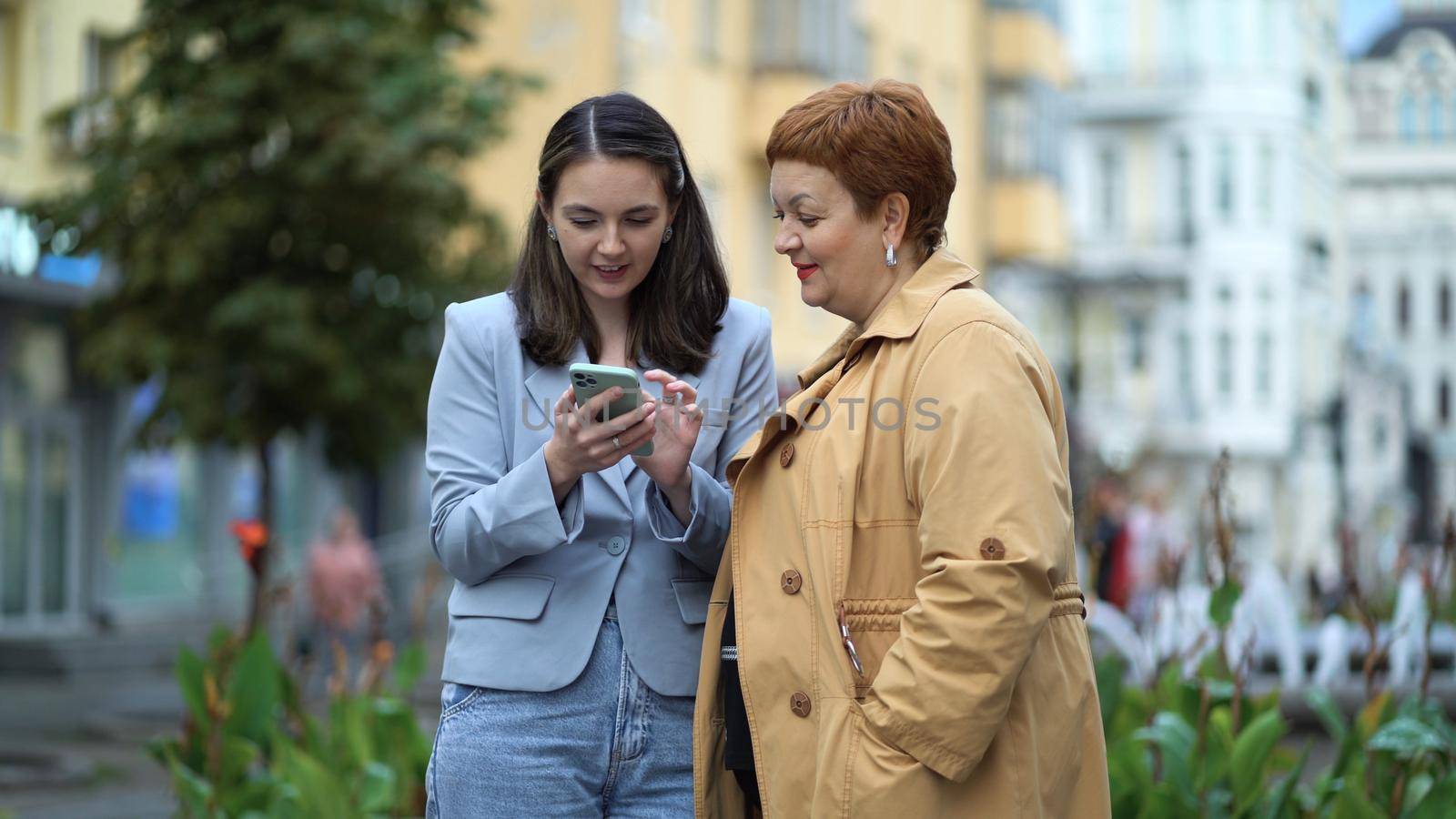 Two women smiling and use phone while standing on the city street. Women enjoy outdoor communication, gossip, online shopping, discuss social media.