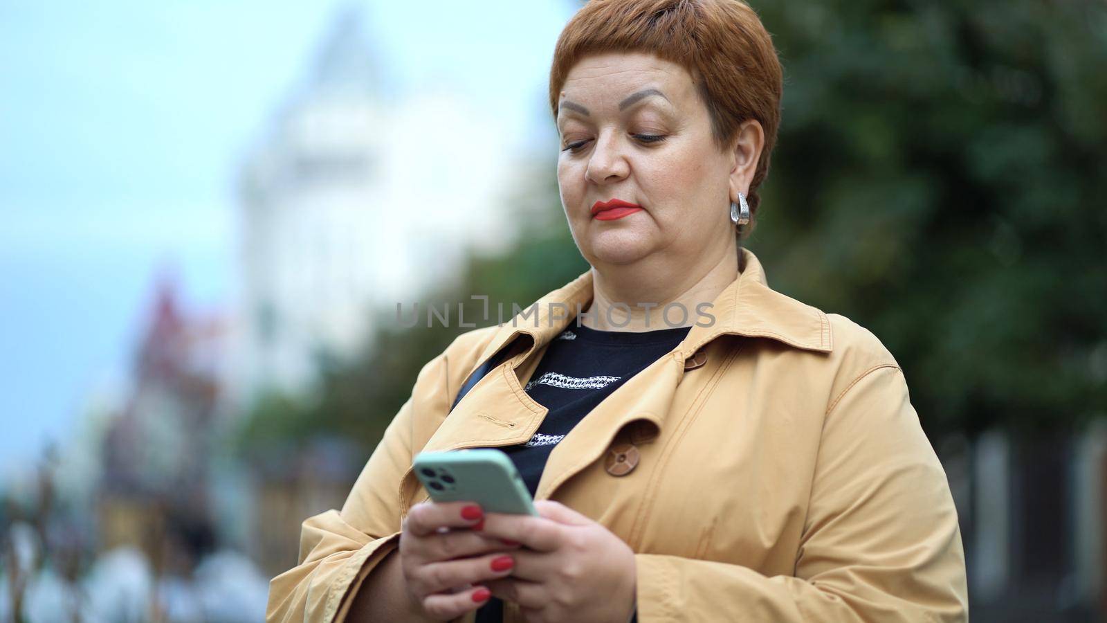 An elegant woman with short red hair and a raincoat is standing on the street and using a smartphone.