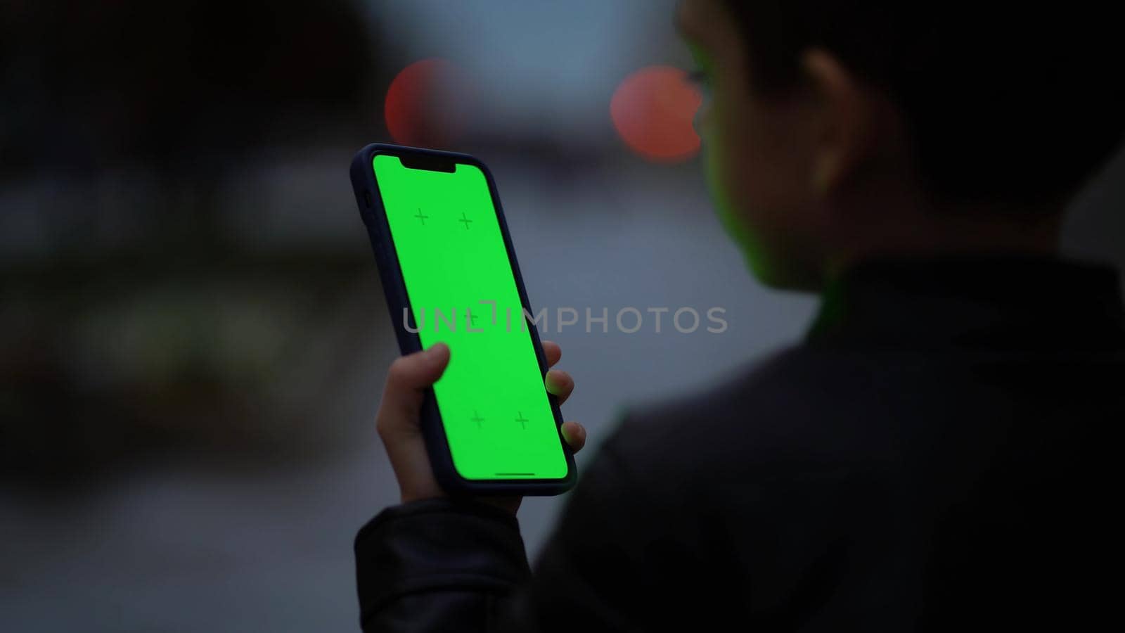 Boy tapping on the green screen of his smartphone. A child with a phone in his hands looks at the chromakey at night in the fall.