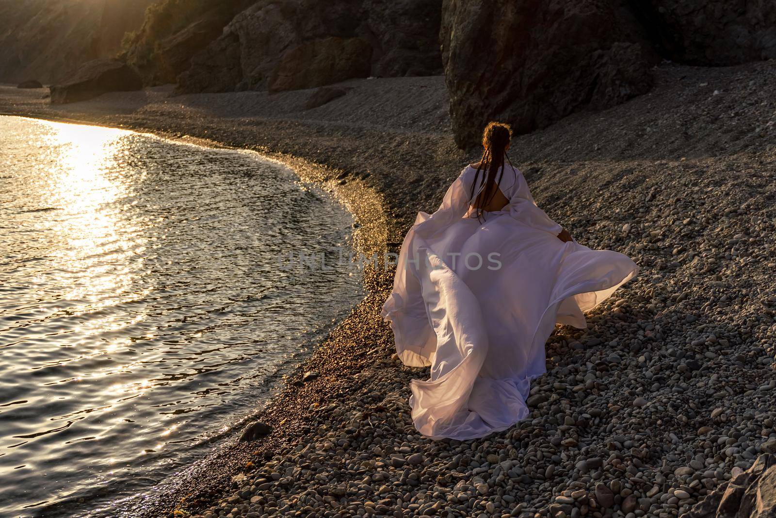 A mysterious female silhouette with long braids stands on the sea beach with mountain views, Sunset rays shine on a woman. Throws up a long white dress, a divine sunset