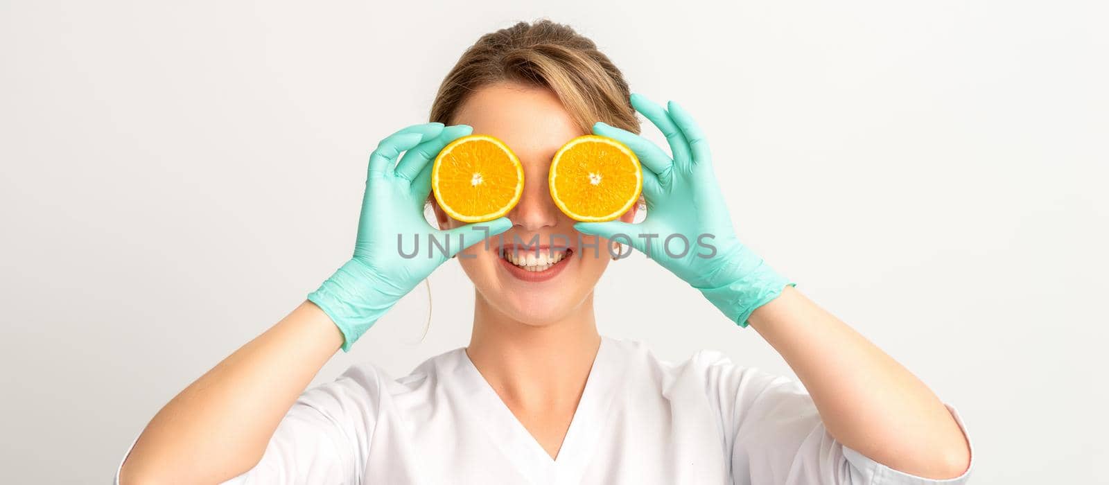 Portrait of young caucasian smiling female beautician covering eye with two orange slices wearing gloves over a white background