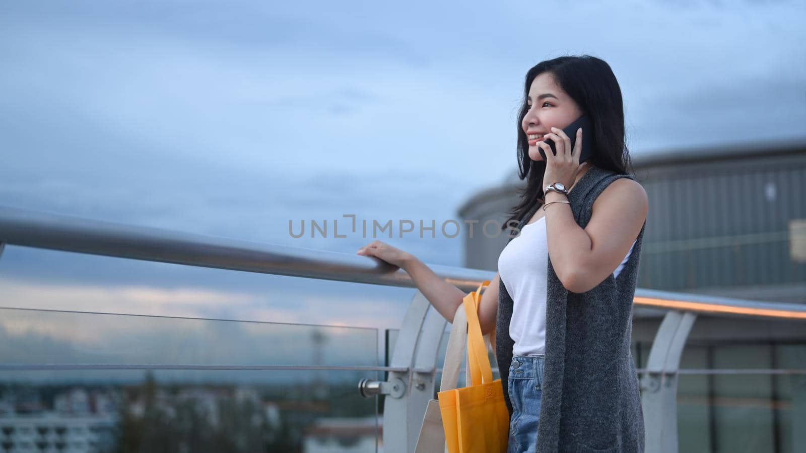 Modern young asian woman talking on mobile phone while standing on rooftop terrace with overlooking the city at sunset view by prathanchorruangsak