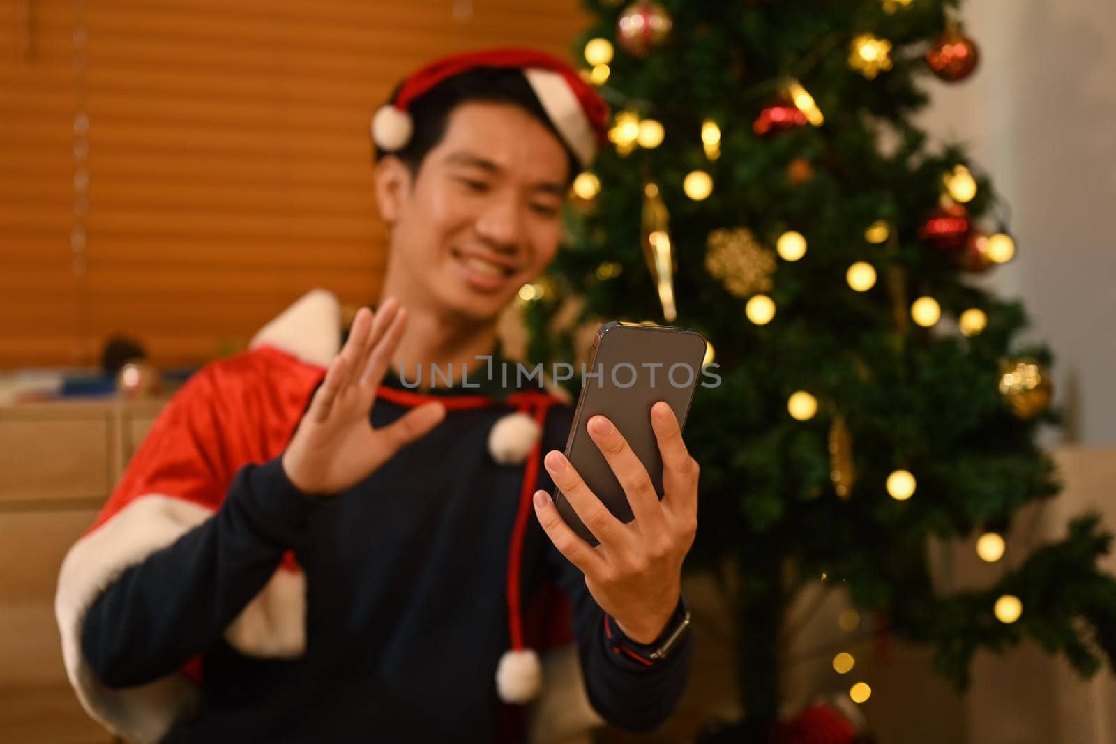Happy man wearing Santa hat making video call on smart phone while sitting in living room with lights garland decorations by prathanchorruangsak
