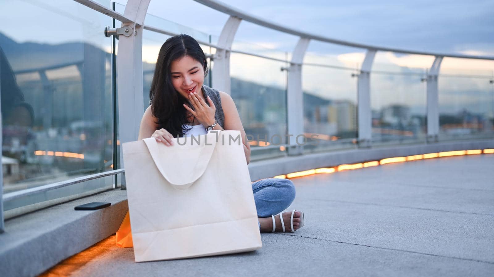 Surprised young woman sitting with shopping bags on rooftop terrace overlooking the city at sunset view in background by prathanchorruangsak