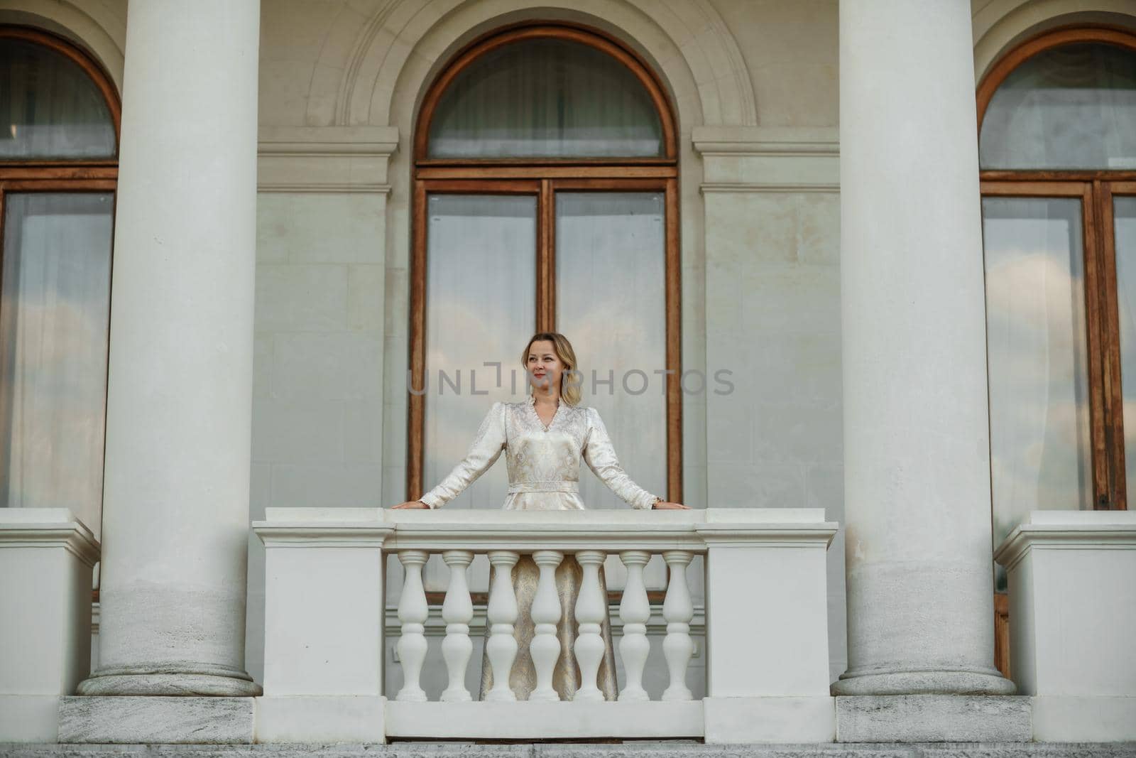 a beautiful mature woman in a long white dress stands on the balcony and looks into the distance.