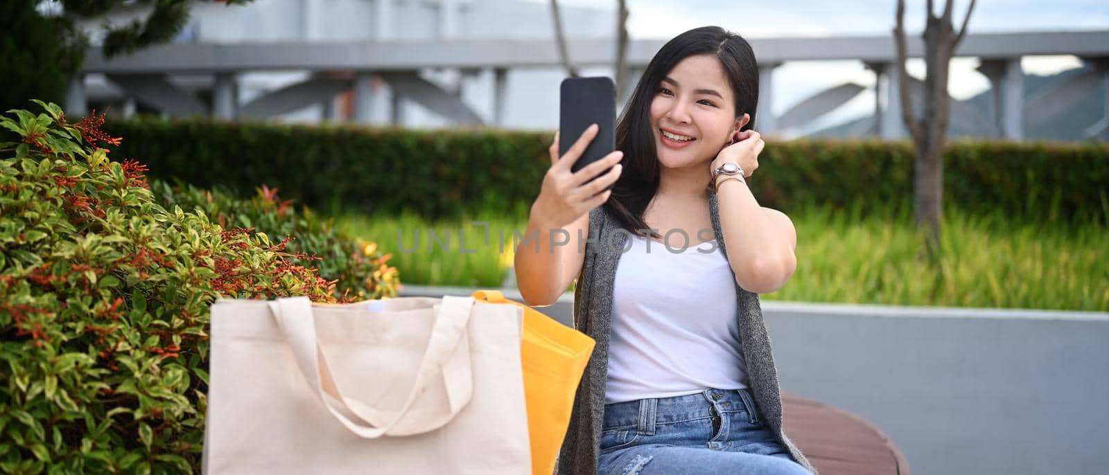 Cheerful young asian sitting on a park bench with shopping bags and takes a selfie on smart phone by prathanchorruangsak