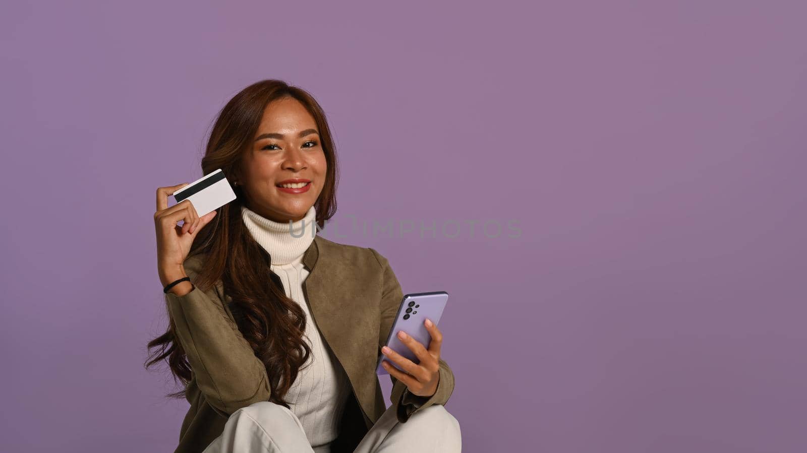 Studio photo of smiling young woman holding mobile phone and credit card sitting on purple background.