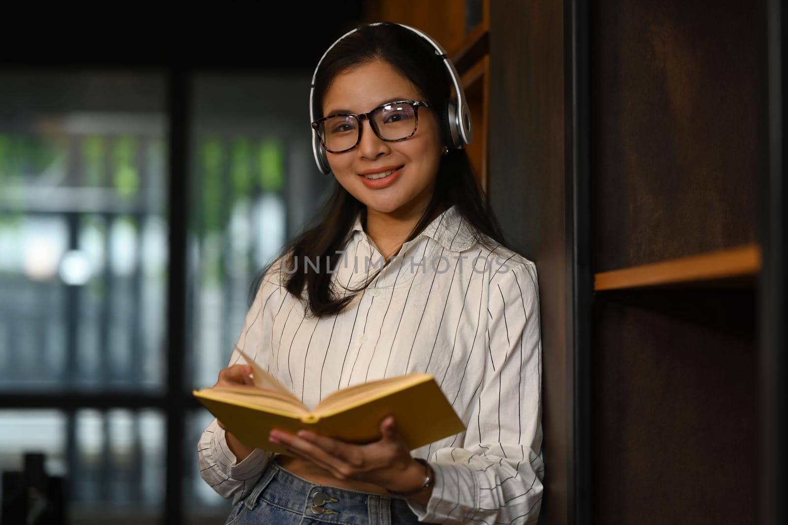 Positive female student reading a book between library bookshelves. Education and people concept by prathanchorruangsak