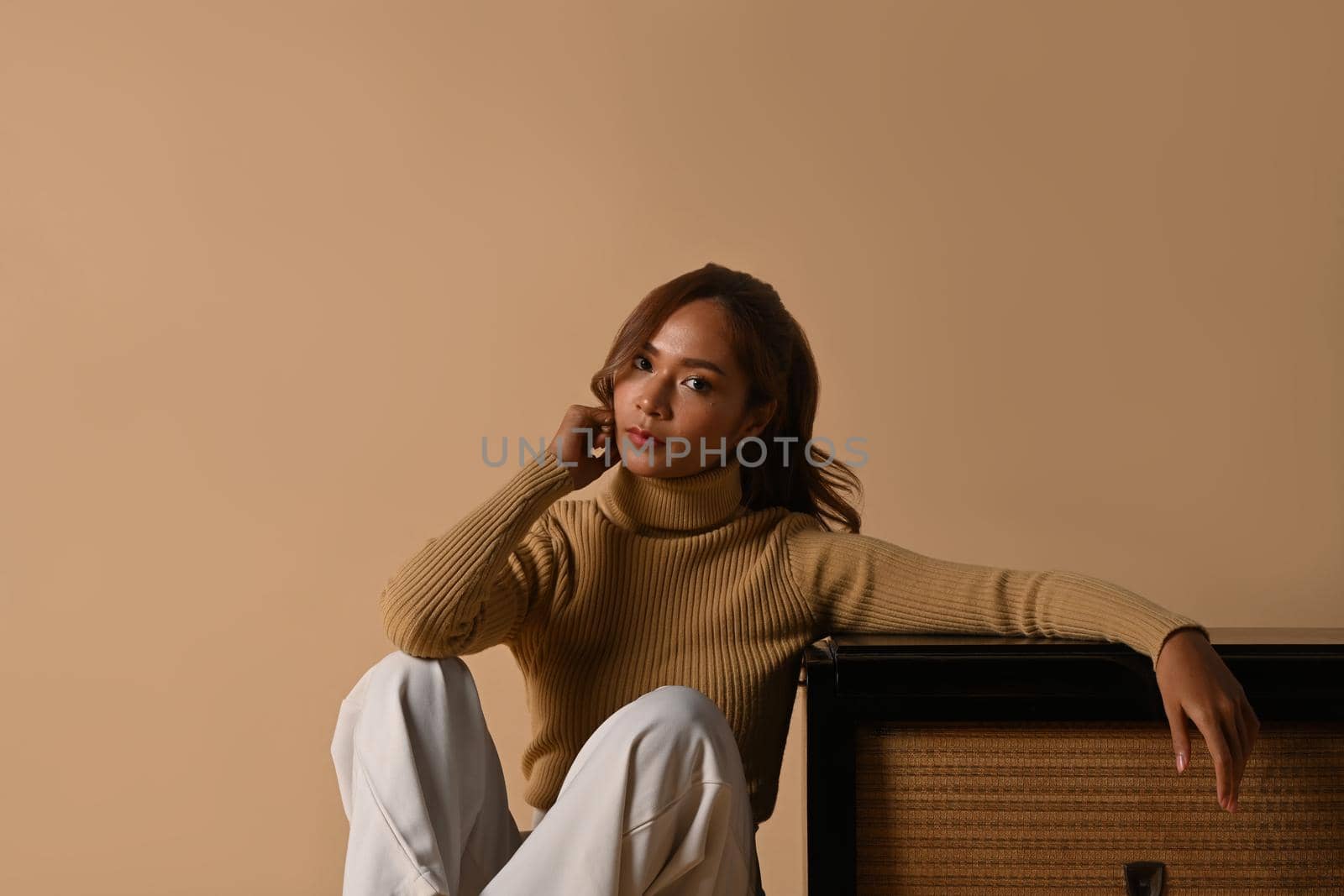 Portrait of fashionable woman wearing trendy sweater sitting near vintage radio, studio shot on beige background by prathanchorruangsak