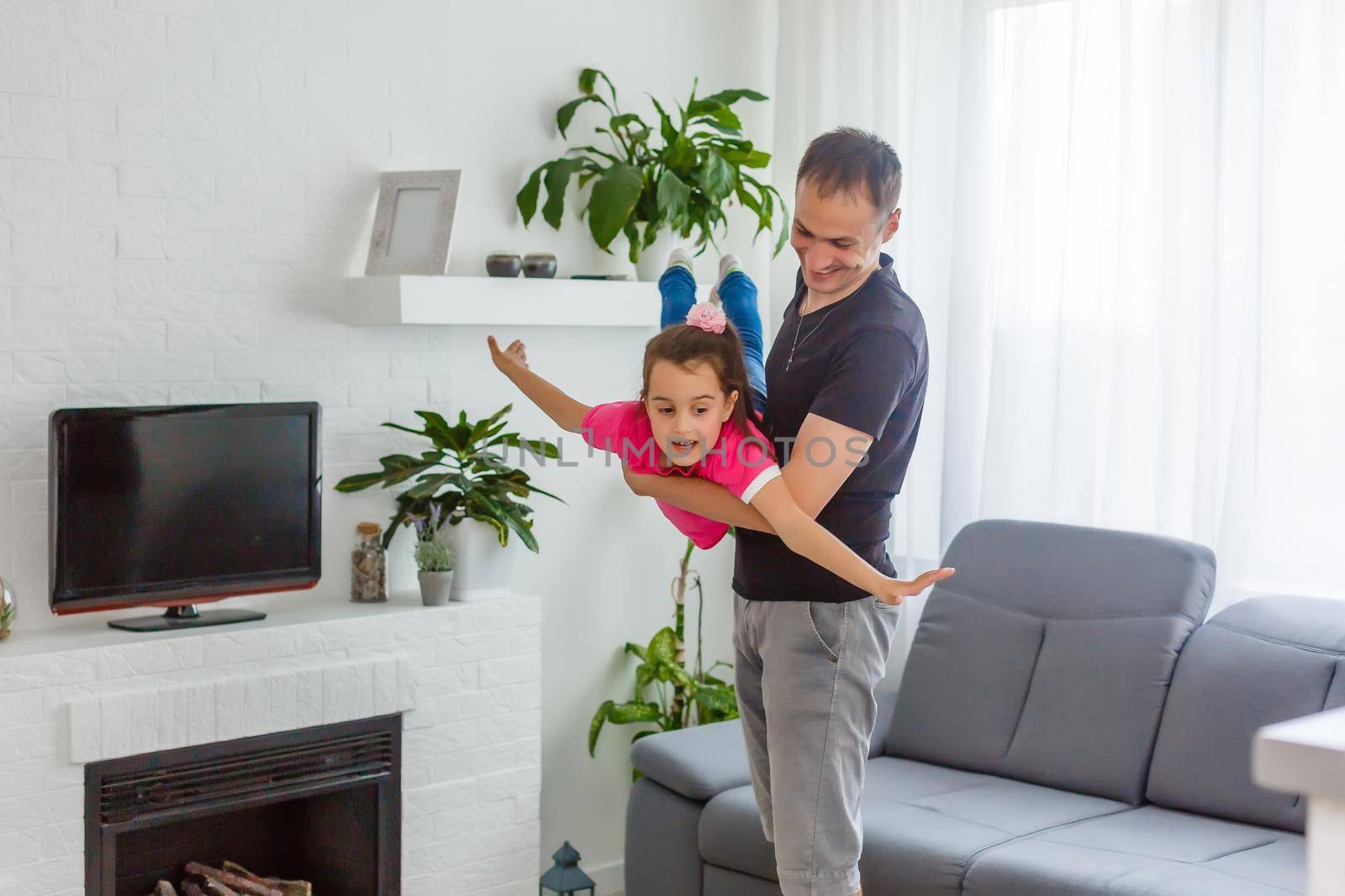 I can fly. Happy dad is lifting up his little daughter while standing in living room.