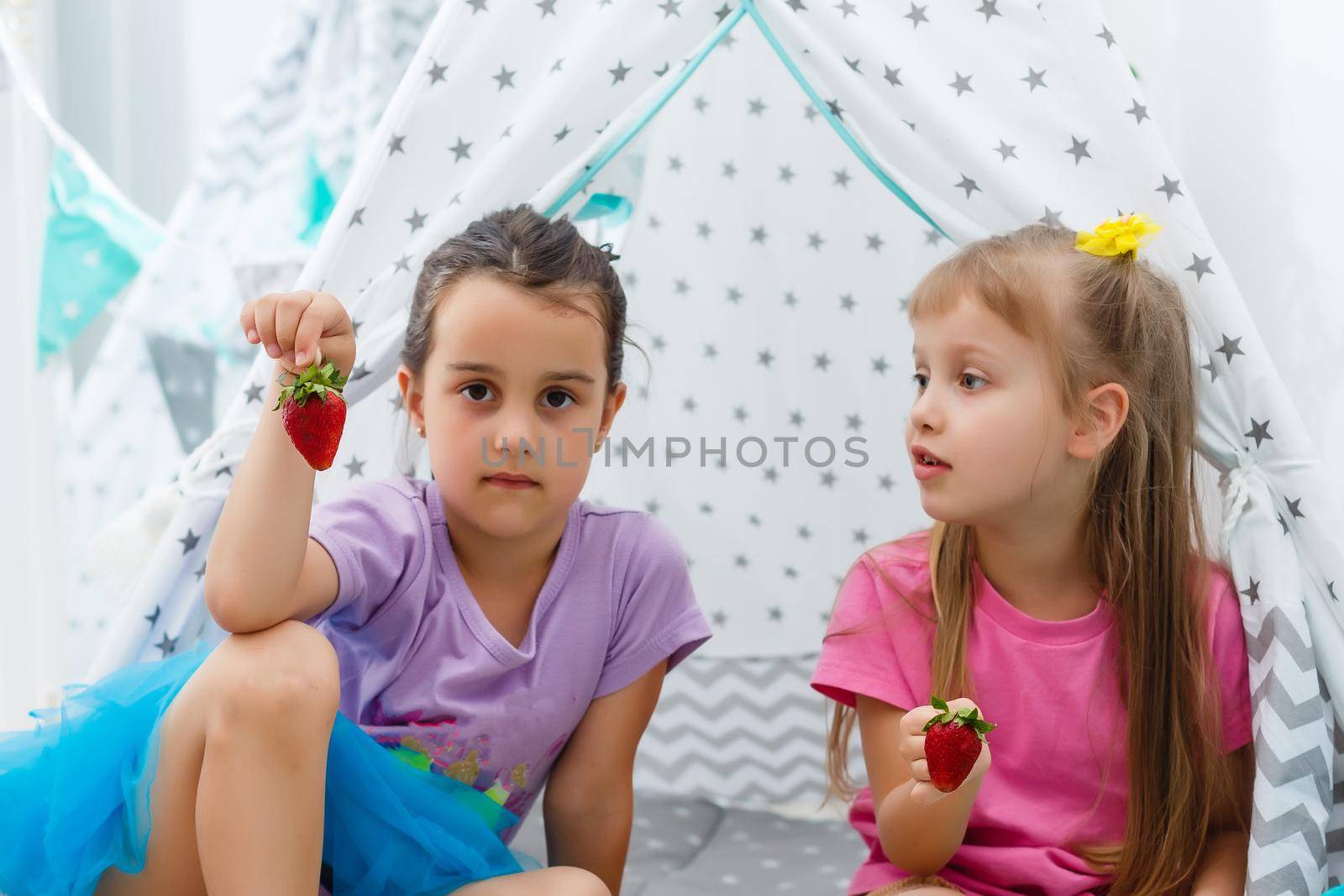 two little girls eat strawberries at home near wigwam tent