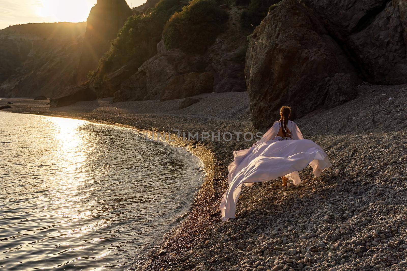 A mysterious female silhouette with long braids stands on the sea beach with mountain views, Sunset rays shine on a woman. Throws up a long white dress, a divine sunset. by Matiunina